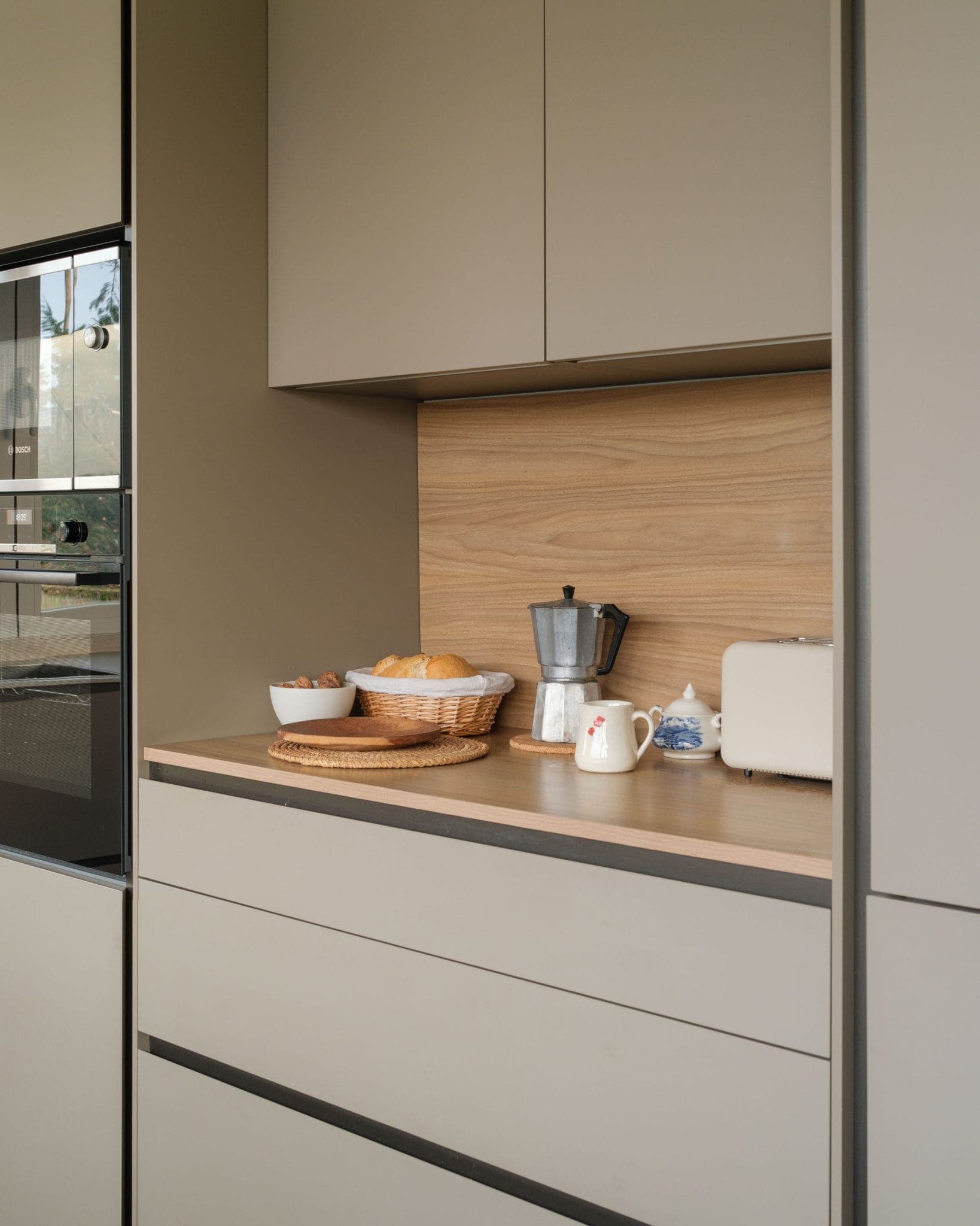Modern kitchen with beige cupboards and wooden credenza. The worktop houses a woven basket containing bread, a bowl of fruit, an Italian coffee maker, a white toaster, a small blue and white teapot and a milk jug. An oven is integrated into the cupboards.
