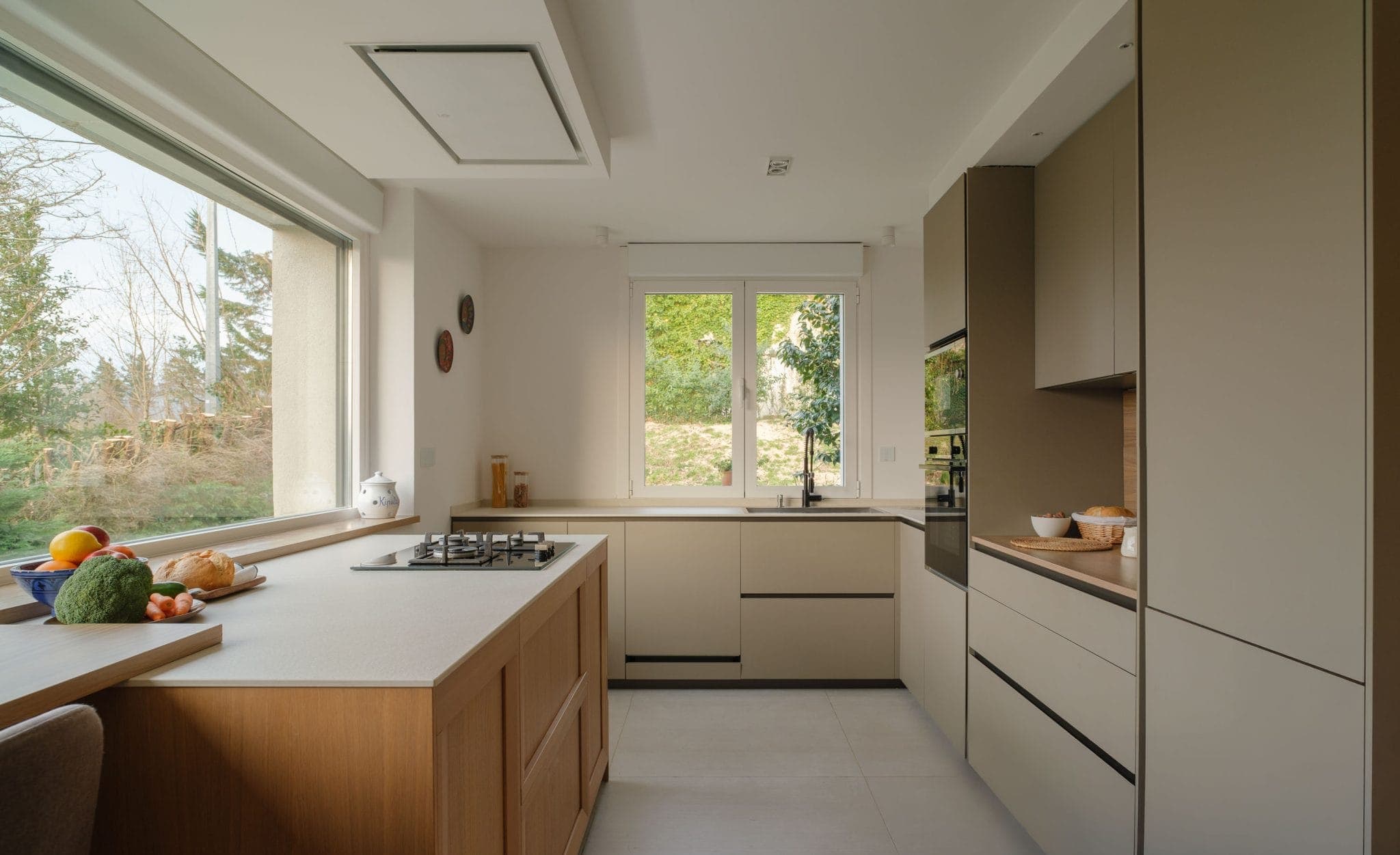 Modern kitchen with light-colored cabinets and countertops. A large window overlooking the trees lets in natural light. A hob and a variety of vegetables sit on the countertop. Neutral tones create a clean, airy atmosphere.