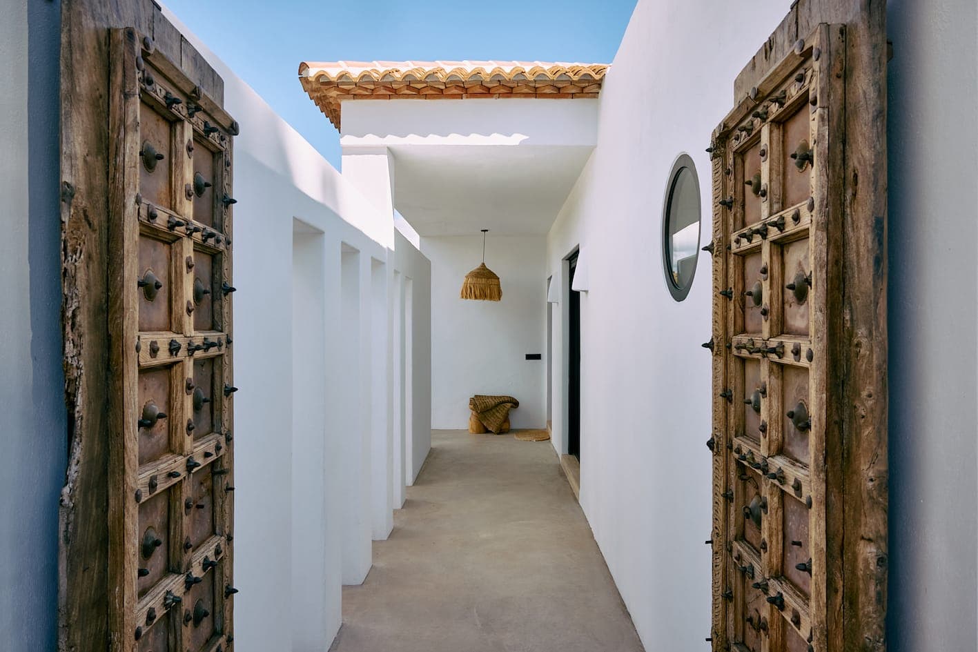 A narrow corridor with white walls and a tiled roof is framed by elaborate wooden doors. Natural light casts shadows along the passage. At the far end, a wicker chair and a hanging lamp can be seen. A round window adorns the right-hand wall.