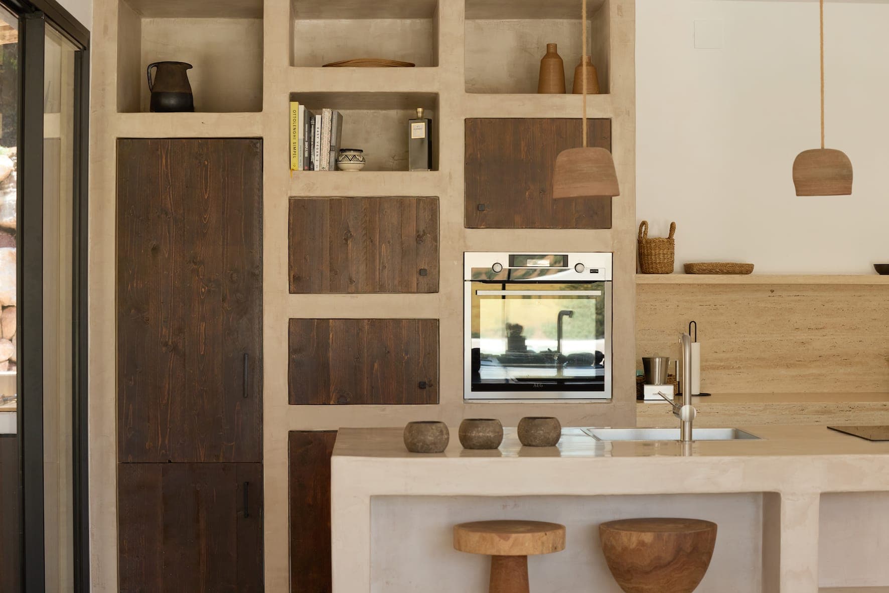 The modern, rustic kitchen features wooden cabinets, a built-in oven, pendant lights and a worktop with stools. Open shelves house books and jars, while decorative objects in natural tones embellish the space.