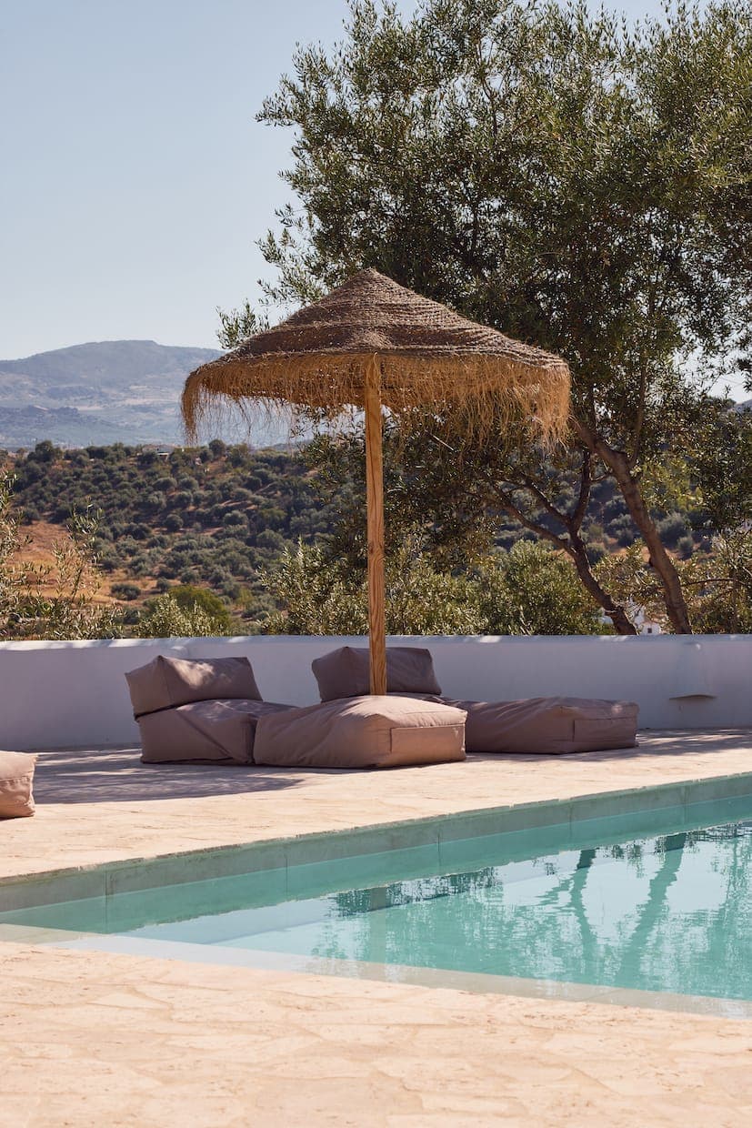 A peaceful poolside landscape, with straw parasol and brown cushions, amidst olive trees. The clear sky and hills in the distance evoke a serene, sunny day in a Mediterranean setting.