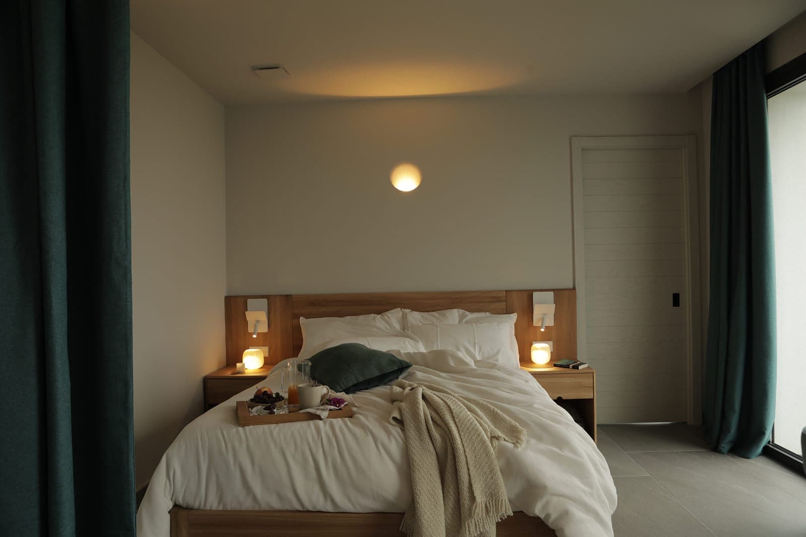 A minimalist bedroom with a wooden bed, soft white linen and a beige plaid. Two lit bedside lamps, a wall sconce above the bed and a breakfast tray add a touch of warmth. Dark curtains partially conceal a large window.