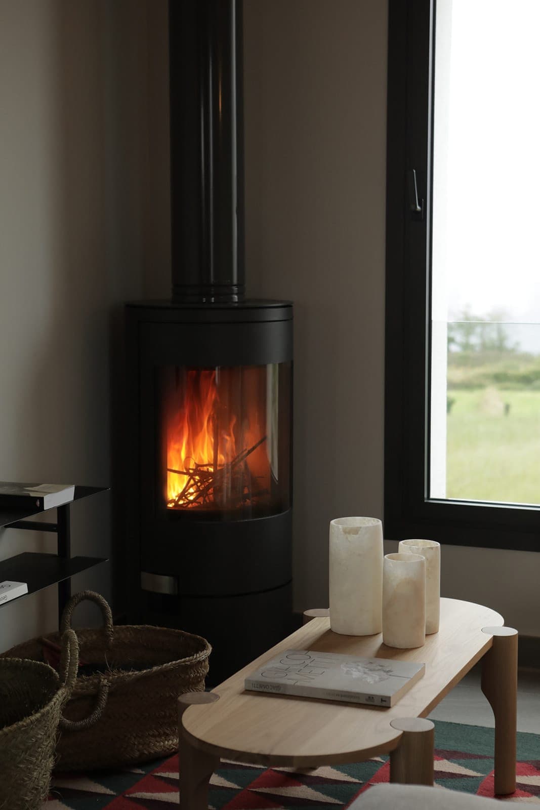 A cozy living room features a modern black wood-burning stove with a visible fire. Next to it, a large window offers a view of the greenery. A wooden coffee table holds white candles and a book, while woven baskets sit on a patterned rug.