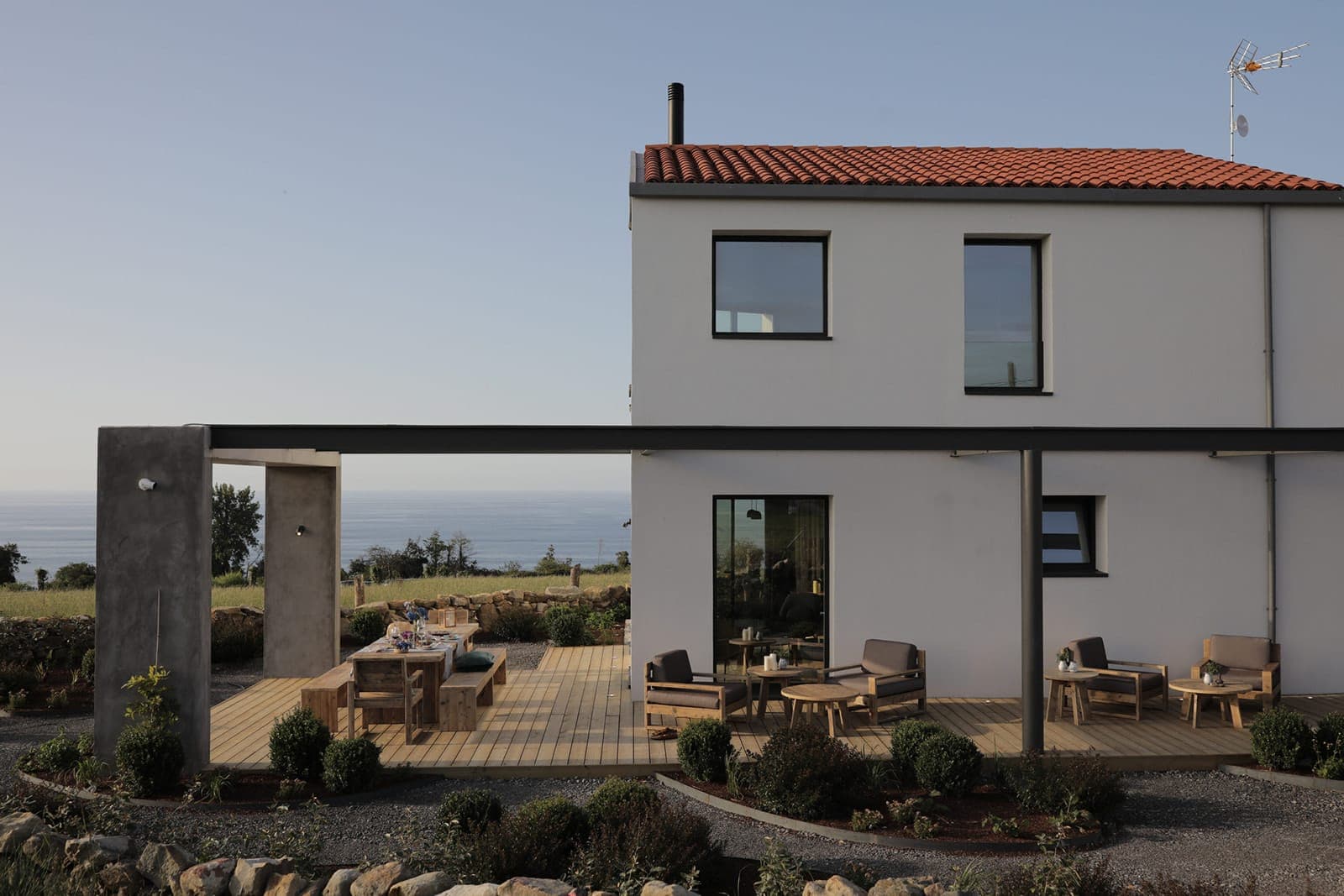 Modern two-story house with a red-tiled roof and white exterior, featuring a wooden deck with outdoor seating and dining area. The house overlooks a grassy yard and the sea in the background under a clear blue sky.