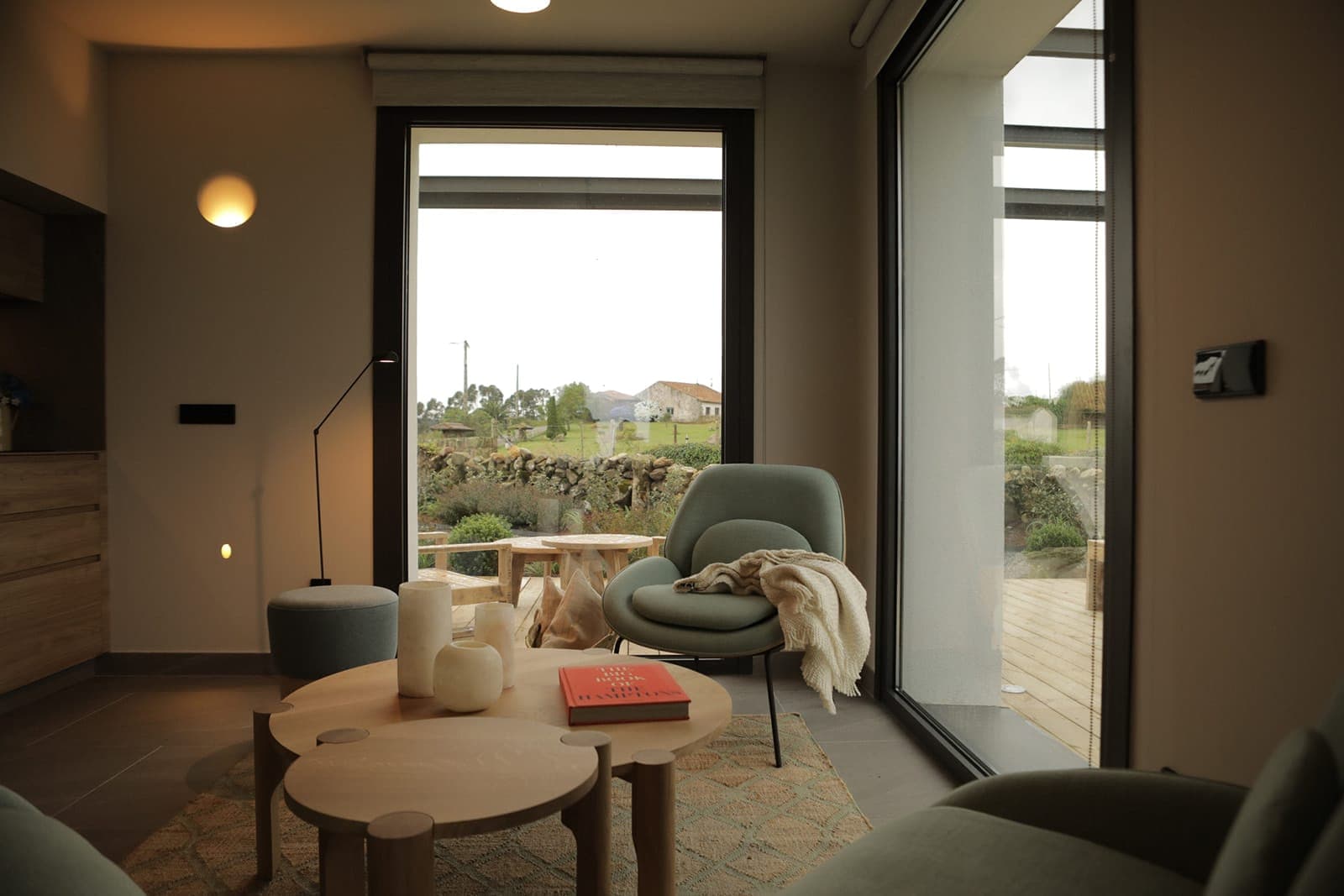 A cosy living room with large windows offering a view of the outside. The room is furnished with a comfortable green armchair with a blanket, round wooden tables adorned with decorative objects and subdued lighting. The atmosphere is warm and welcoming.