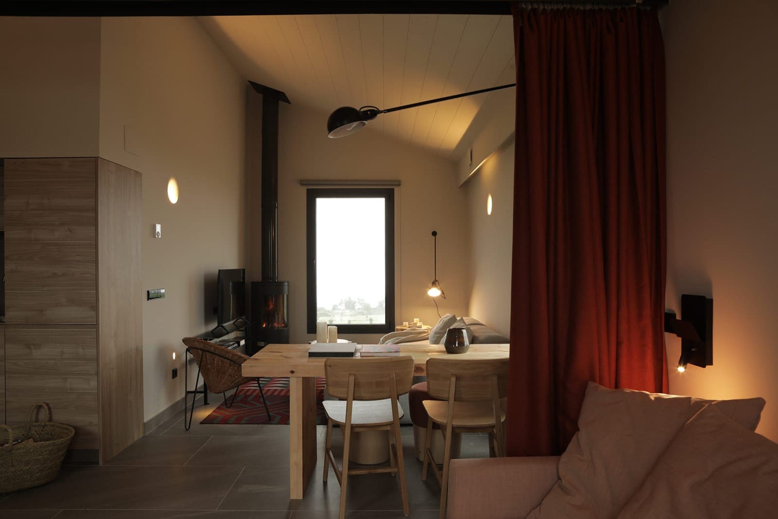Warm, modern-design living space with paneled interior, wood-burning stove, dining table with chairs and beige sofa. A large window lets in natural light, and curtains add a warm touch to the ambience.