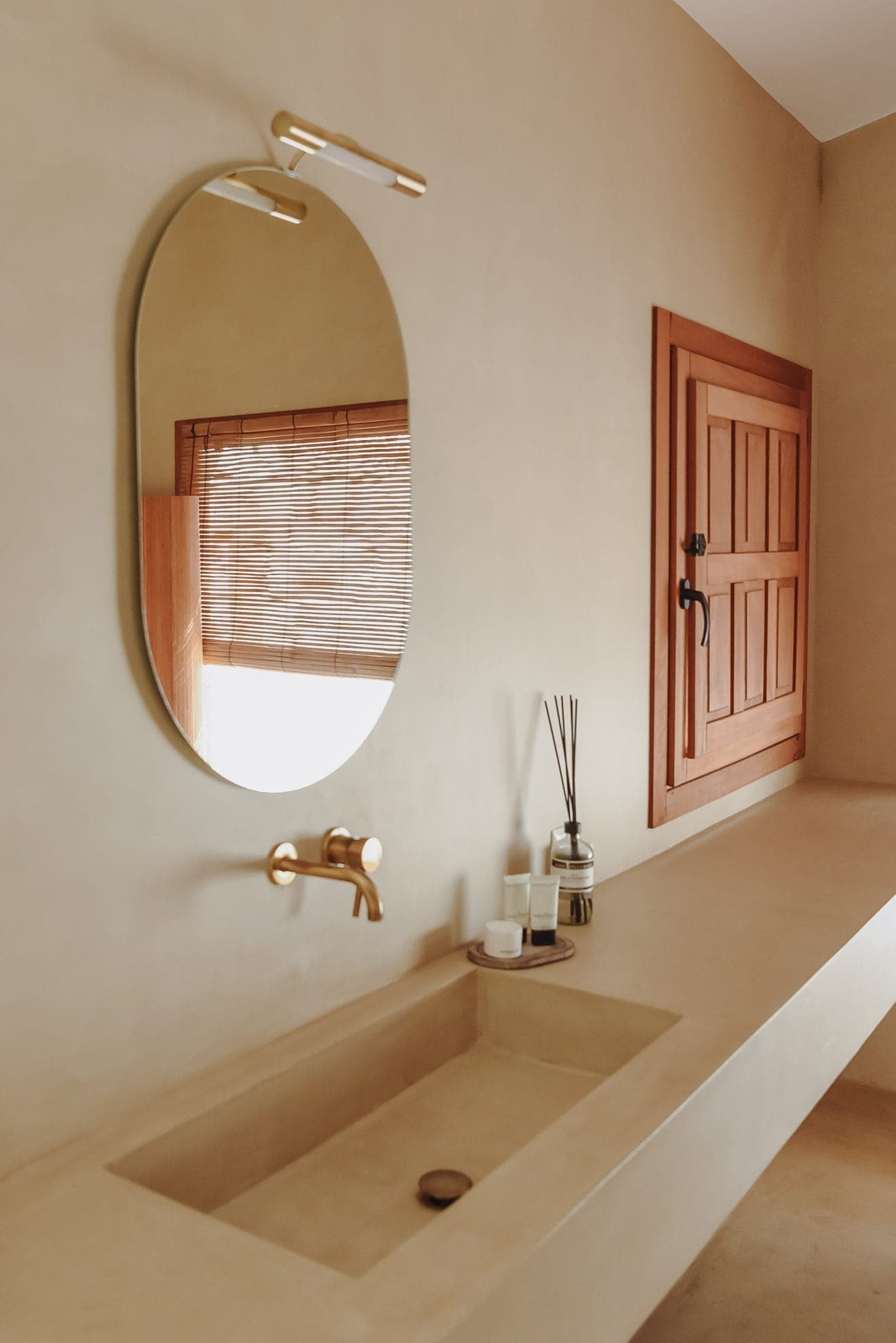 Minimalist bathroom with beige countertop and integrated basin. A hanging oval mirror reflects a window with bamboo blinds. A wooden cupboard door hangs on the wall, and a set of toiletries and a perfume diffuser are neatly arranged nearby.