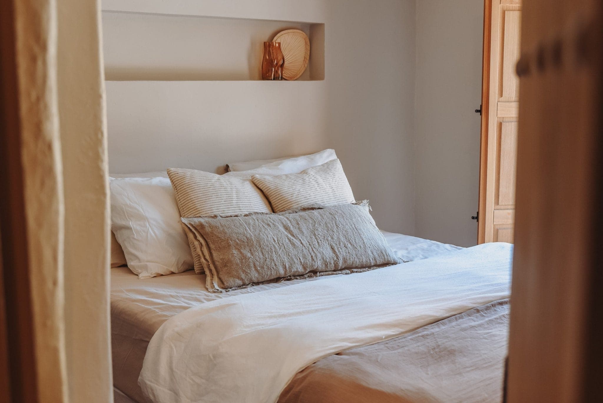 A cozy room with a carefully made bed, embellished with white and beige sheets and pillows. Above the bed is a small built-in shelf with decorative objects. Warm, natural light from a window gently illuminates the space.