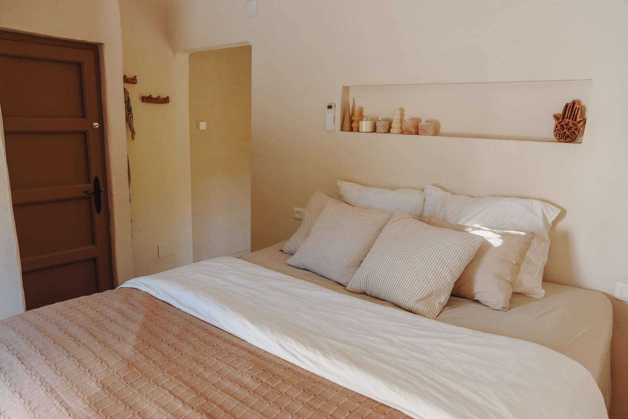 A cozy bedroom with a carefully made bed, complemented by beige and cream pillows and a quilted blanket. A shelf above the bed houses decorative objects. The walls and door are in warm, natural tones, creating a serene atmosphere.