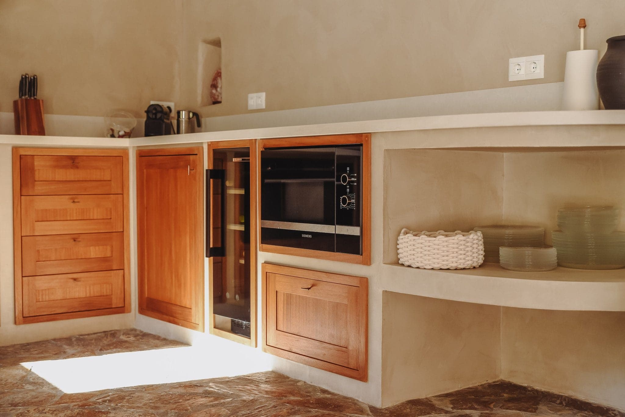 A minimalist kitchen with light wood cabinets, a built-in oven and open shelves housing plates and baskets. The worktop houses a knife block and small appliances. The floor is tiled with stone and the walls are a neutral beige.