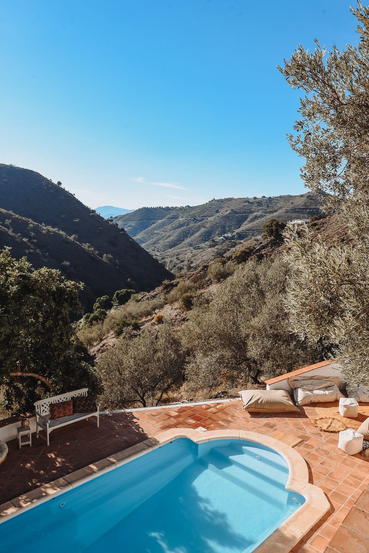 Vue panoramique d'un jardin avec une petite piscine rectangulaire et des carreaux en terre cuite, surplombant un paysage montagneux. Un banc blanc et des coussins sont installés sous un arbre, avec des collines ondulantes et un ciel bleu clair en arrière-plan.
