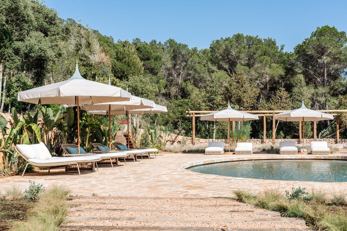 Outdoor pool area with several white parasols and sun loungers on a stone terrace. The pool is surrounded by lush vegetation and trees. The clear, sunny sky reinforces the peaceful atmosphere.
