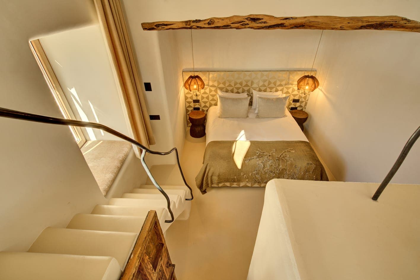 A cosy, minimalist bedroom seen from a staircase. It features a carefully designed double bed, two hanging lamps, a wooden beam and a textured blanket. Warm lighting creates a welcoming, serene atmosphere.