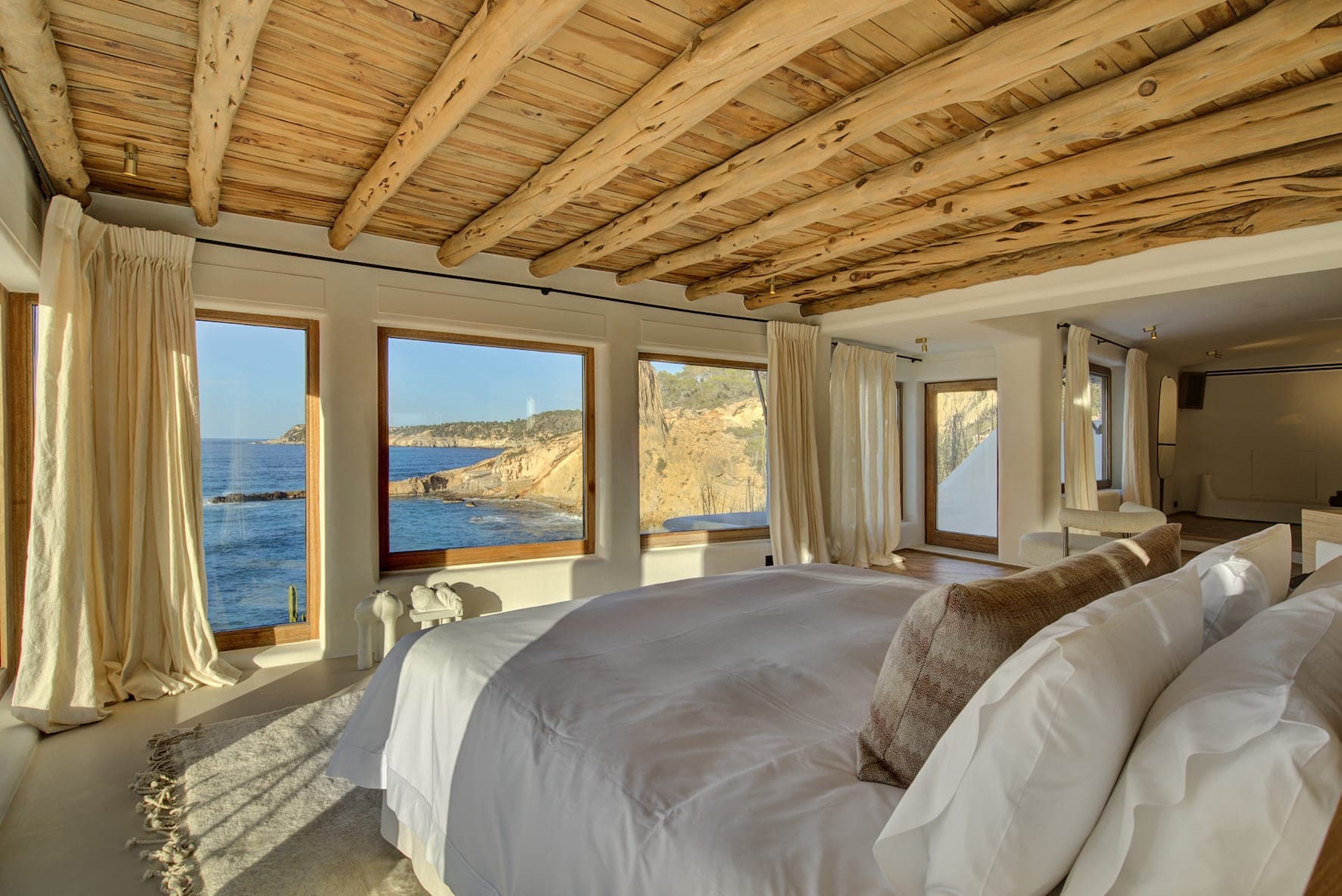 A bedroom with a king-size bed, white linens and plenty of pillows, is surrounded by picture windows offering breathtaking ocean views. Its rustic wood ceiling and light curtains create a warm, welcoming atmosphere.