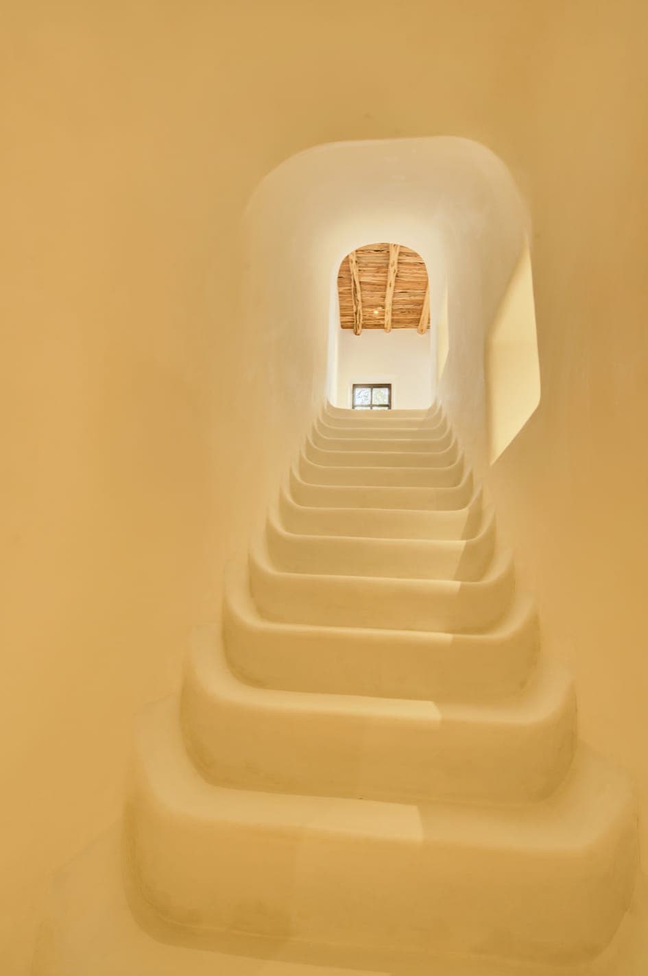 A gently curving white staircase leads up to an arched opening with a small window. Natural light filters in, highlighting the rustic wooden ceiling. The overall effect is minimalist and serene.
