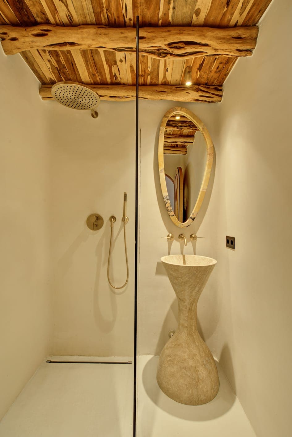 A modern, minimalist bathroom with wooden ceiling and unique stone pedestal sink. It features a round mirror with wooden frame, an elegant glass shower partition and a ceiling-mounted rain shower head. The walls are smooth and bright.