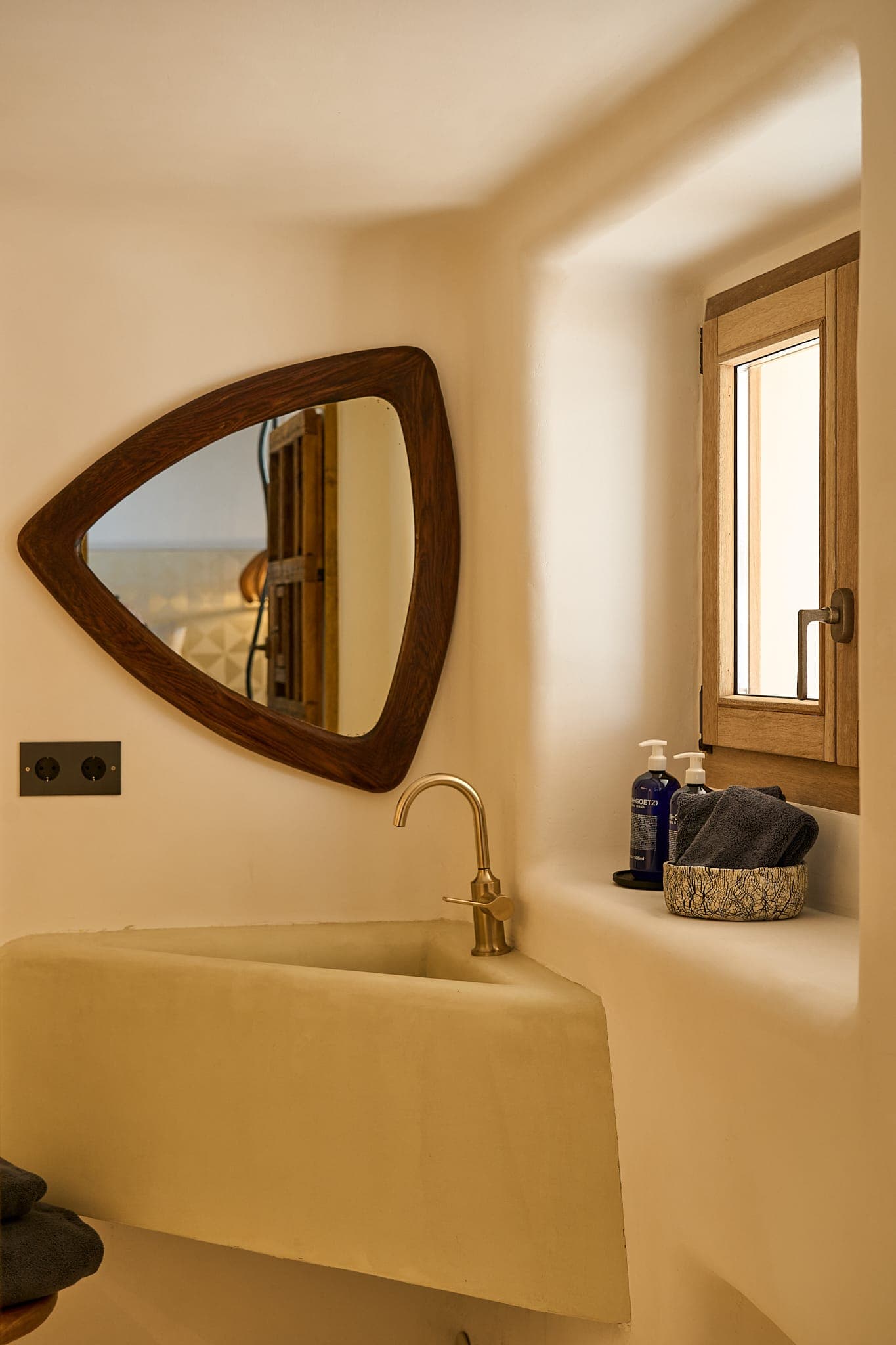 A cosy bathroom corner with a wood-framed triangular mirror above a minimalist washbasin. A brass faucet is installed above the sink. A small window with wood panelling is on the right, and toiletries are placed on the sill. Soft, warm lighting enhances the ambience.