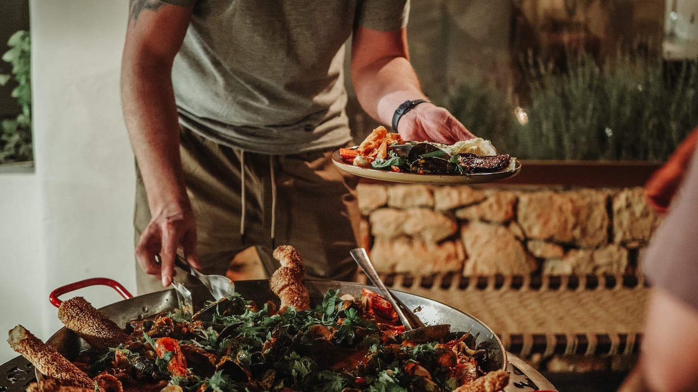 One person serves dishes on a large, colorful plate, garnished with a variety of ingredients, including greens and breadsticks. The setting appears to be that of a casual dining room with rustic decor.