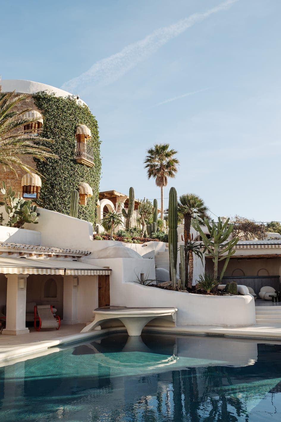 This luxurious Mediterranean-style villa, with its white facade partially covered in ivy and its many balconies, is adorned with large palm trees and a variety of cacti. In the foreground, a large, inviting swimming pool reflects the clear sky.