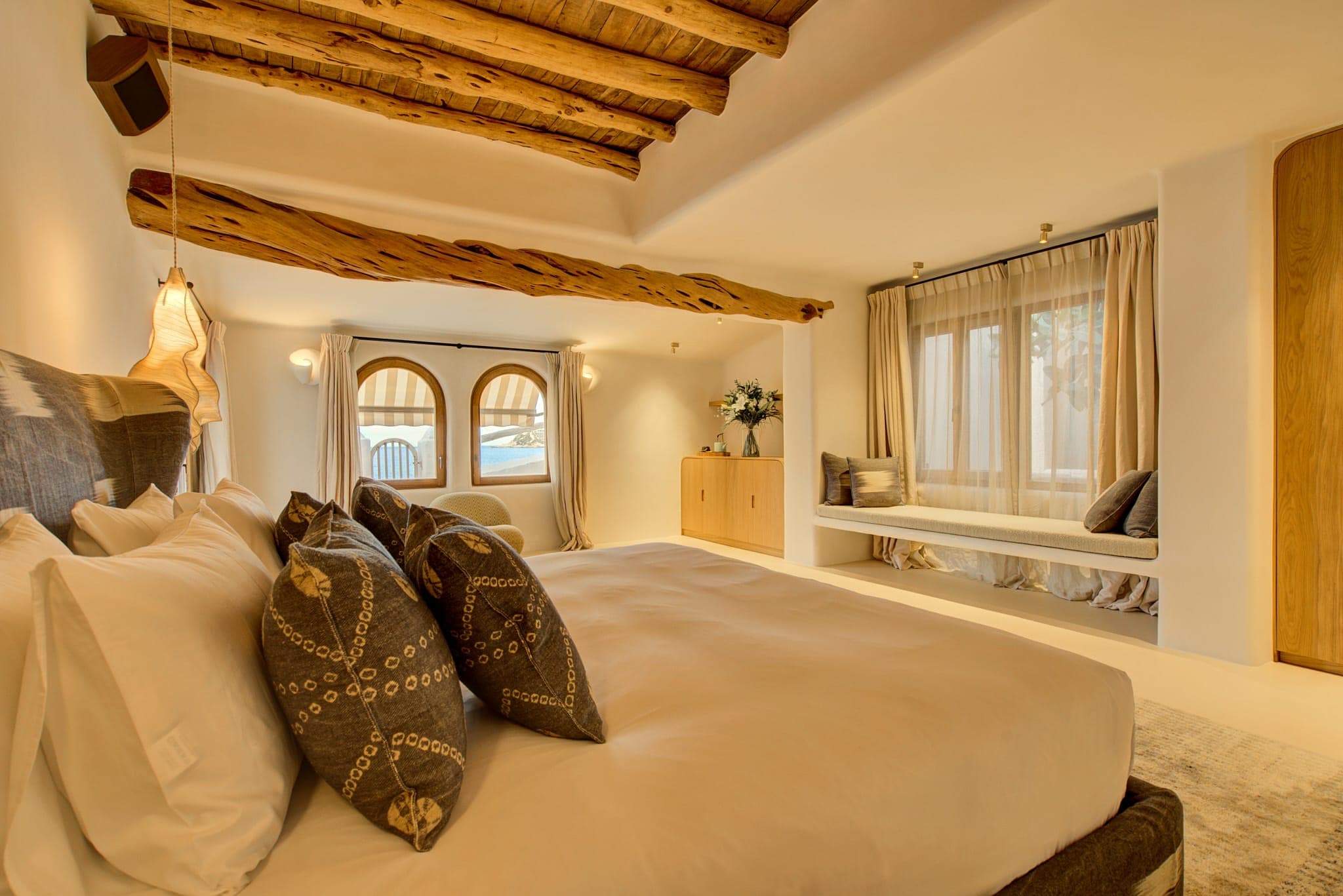 A comfortable bedroom features a king-size bed with patterned pillows, exposed ceiling beams and two arched windows. An upholstered bench is adorned with curtains, as is a wooden wardrobe decorated with flowers. The atmosphere of the room is warm and welcoming.