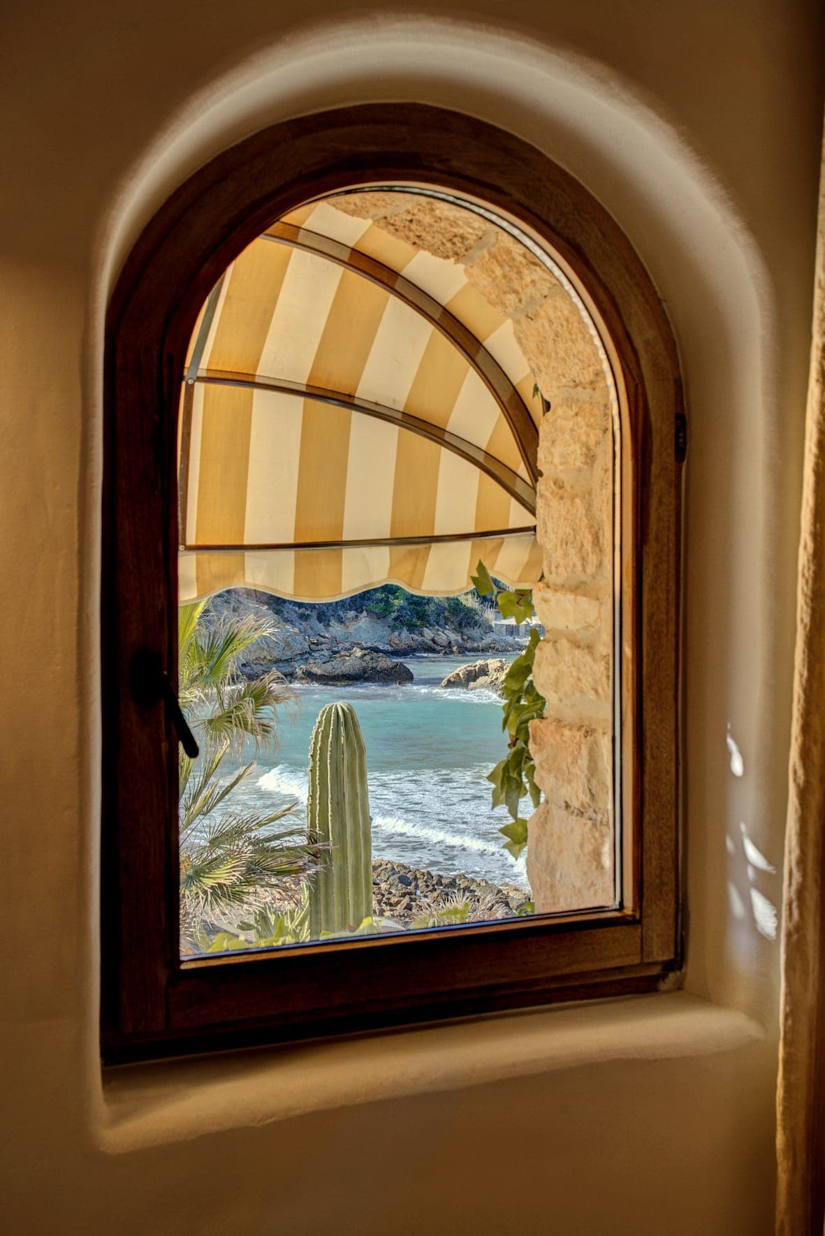 A panoramic view through an arched window with a striped blind reveals a beach with turquoise waters, a rocky shoreline, palm trees and a large cactus. The window is set into a stone wall, creating a rustic setting for this peaceful seaside landscape.
