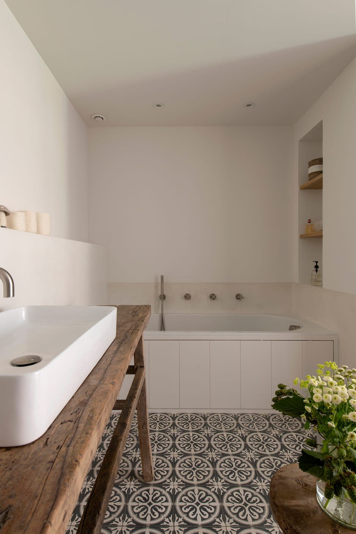 A modern bathroom with a patterned tiled floor, a rustic wooden vanity with a rectangular white sink, a bathtub set into a corner and a vase of white flowers on a small table. The walls are sober and clear.