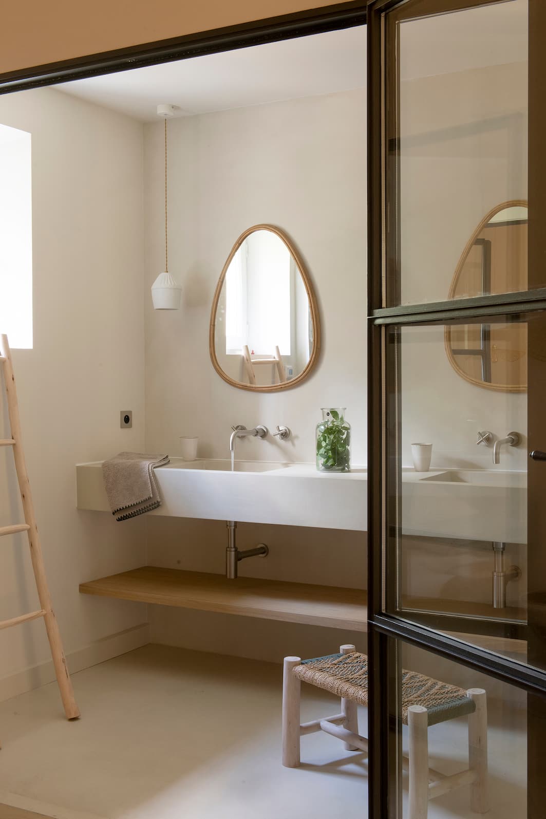 A minimalist bathroom with a large white sink, a wood-framed oval mirror, a hanging lamp, a wooden ladder, a small woven stool and a glass door. A green plant stands near the sink.
