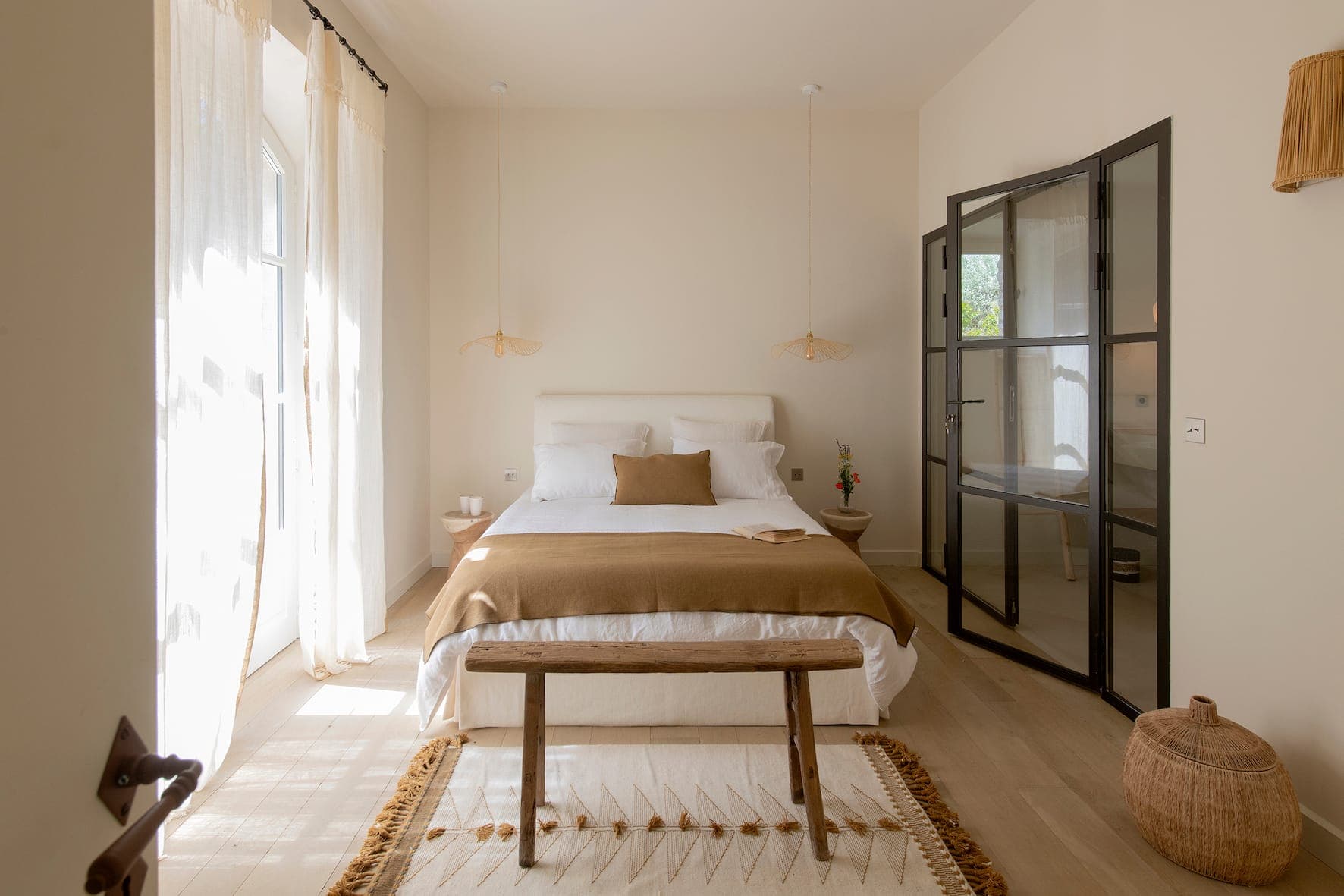 Minimalist bedroom with king-size bed, neutral bedding, wooden bench at foot, woven carpet, hanging lights, sheer curtains, glass doors and natural decor creating a calm, airy atmosphere.