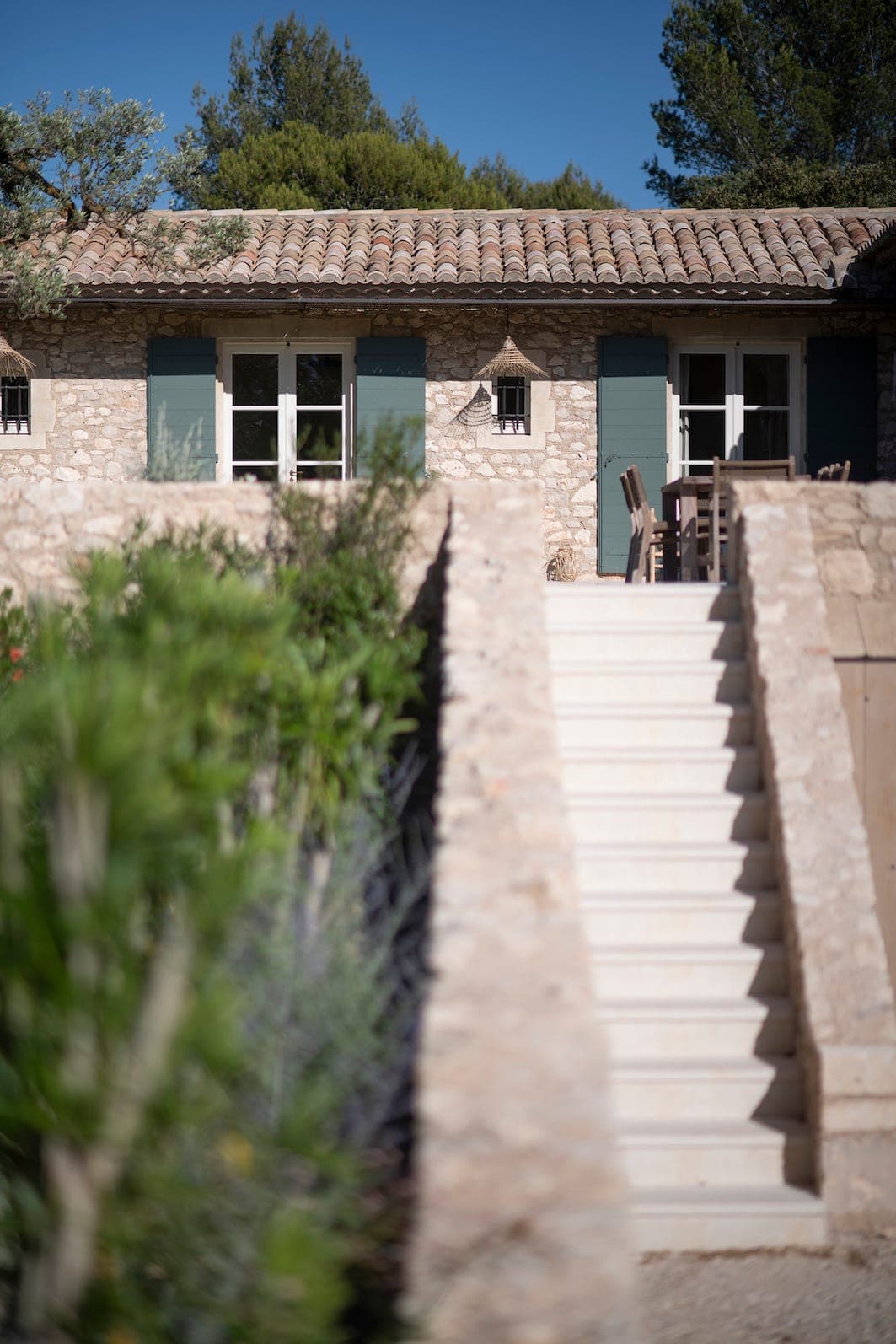 Behind a stone staircase stands a stone house with blue shutters and a tiled roof, surrounded by greenery under an azure-blue sky. Garden furniture is visible near the windows.