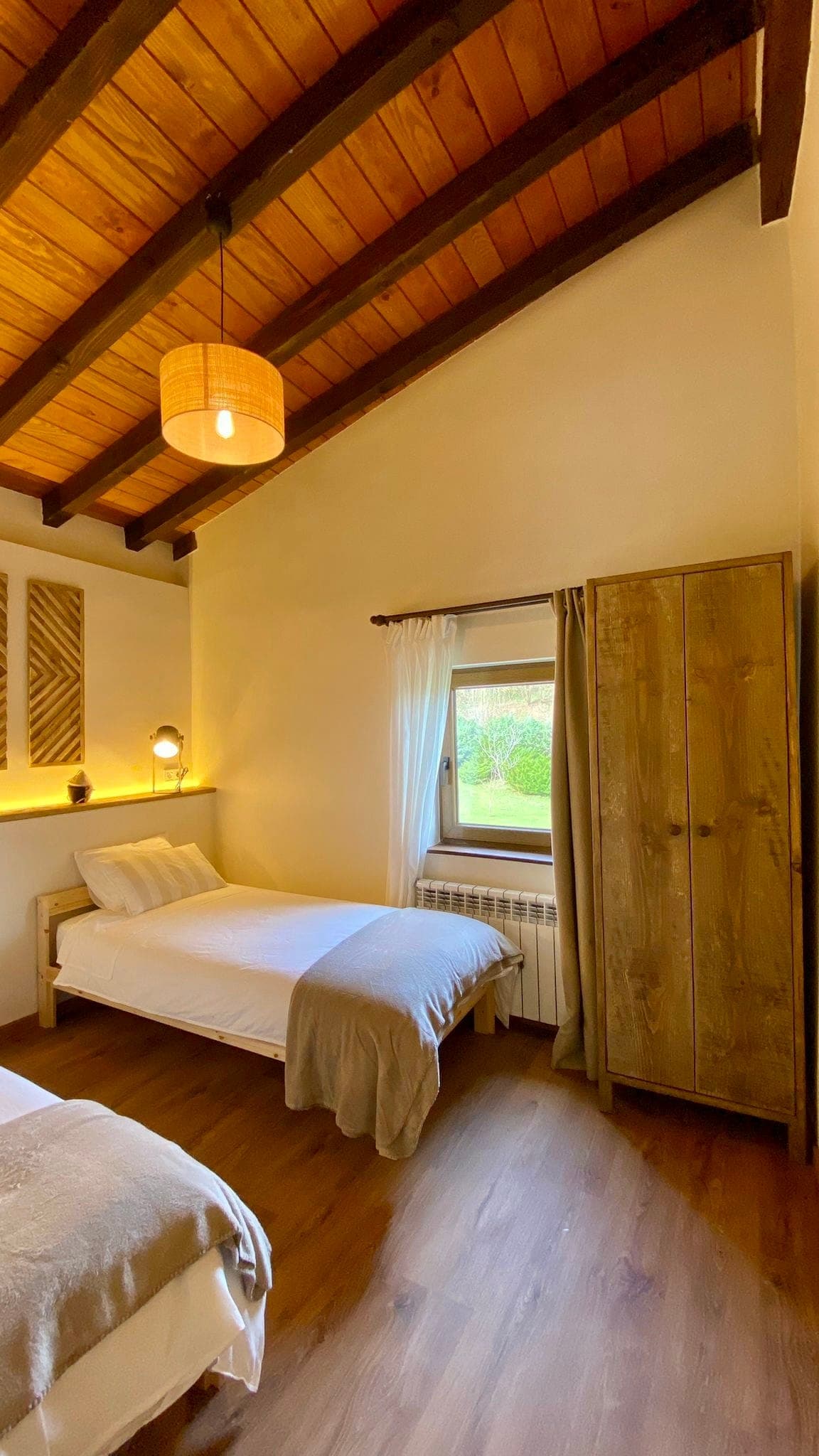 Beamed bedroom with double bed, wardrobe and small window with white curtains. Warm lighting creates a cosy atmosphere. The walls are light and the floor is wood. A wall lamp and works of art hang above the bed.