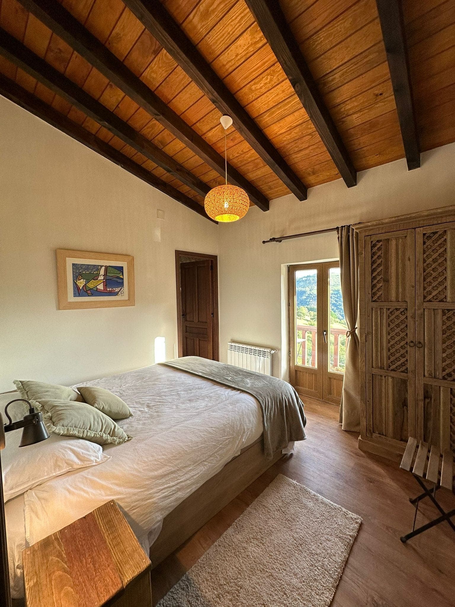 Comfortable room with wooden ceiling and large bed adorned with cushions. A wicker hanging lamp hangs above. Sunlight filters through a window, offering a view of the greenery outside. A wooden wardrobe and a small chair complete the furnishings.