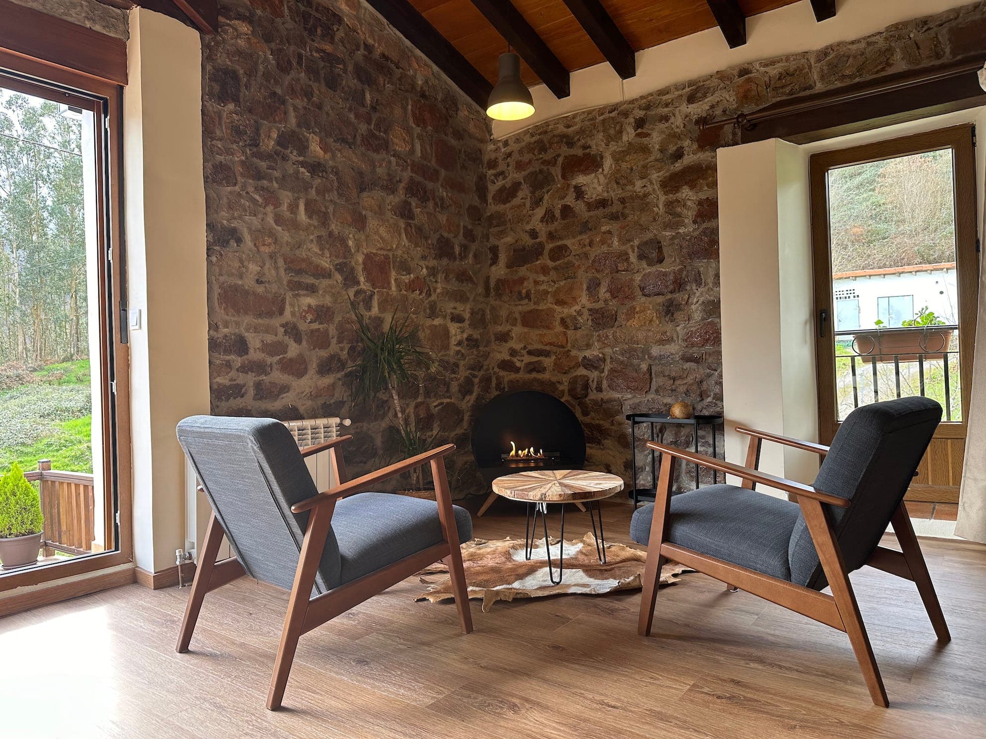 Two modern armchairs face a round marble coffee table set on a cowhide rug, in a warm room with stone walls, parquet flooring, large windows and a small indoor fireplace. Outside, you can see lush vegetation.