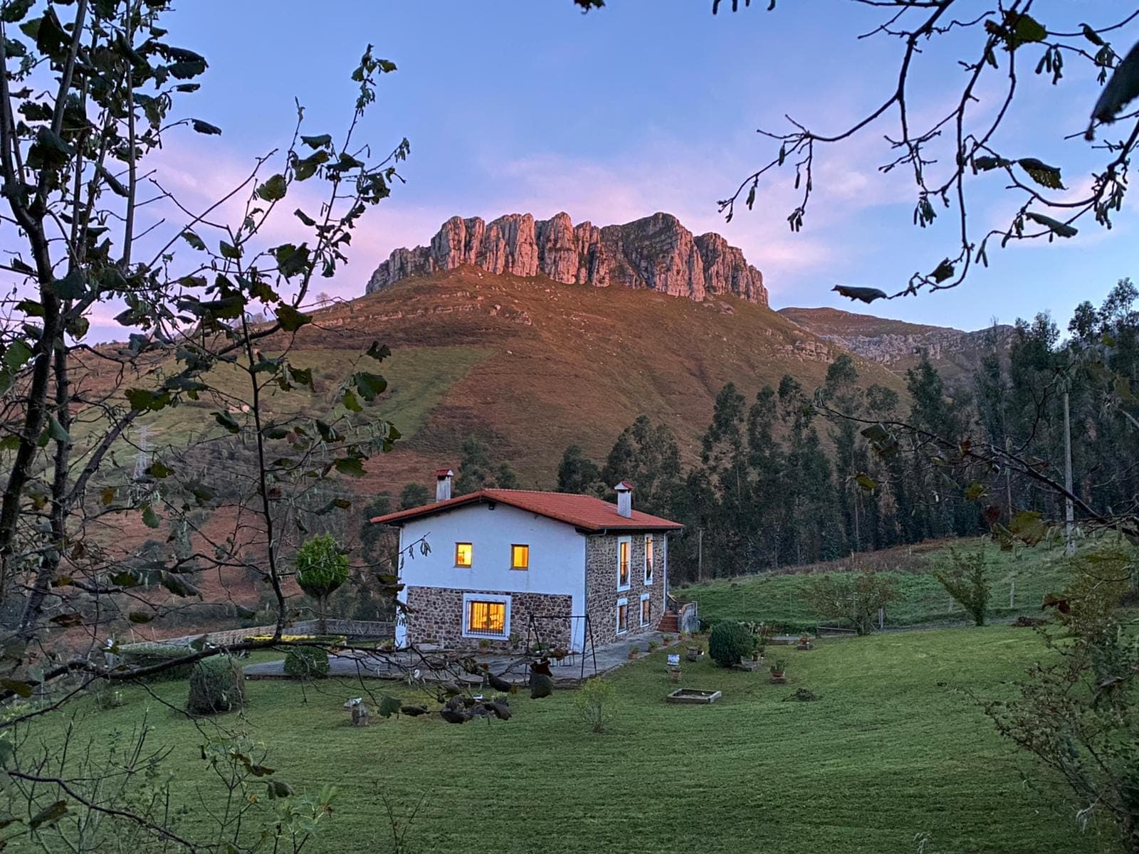 A stone house with bright windows sits on a grassy hill, surrounded by trees, with a rocky mountain peak rising in the background under a pastel pink and blue evening sky.