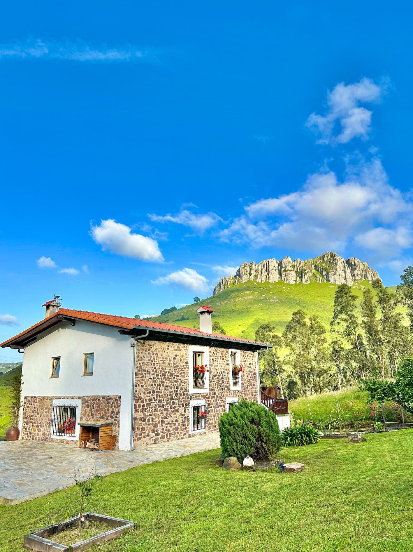 A stone and white house with a red tile roof sits on a grassy lawn, surrounded by green hills and trees, with a rocky cliff and bright blue sky in the background.