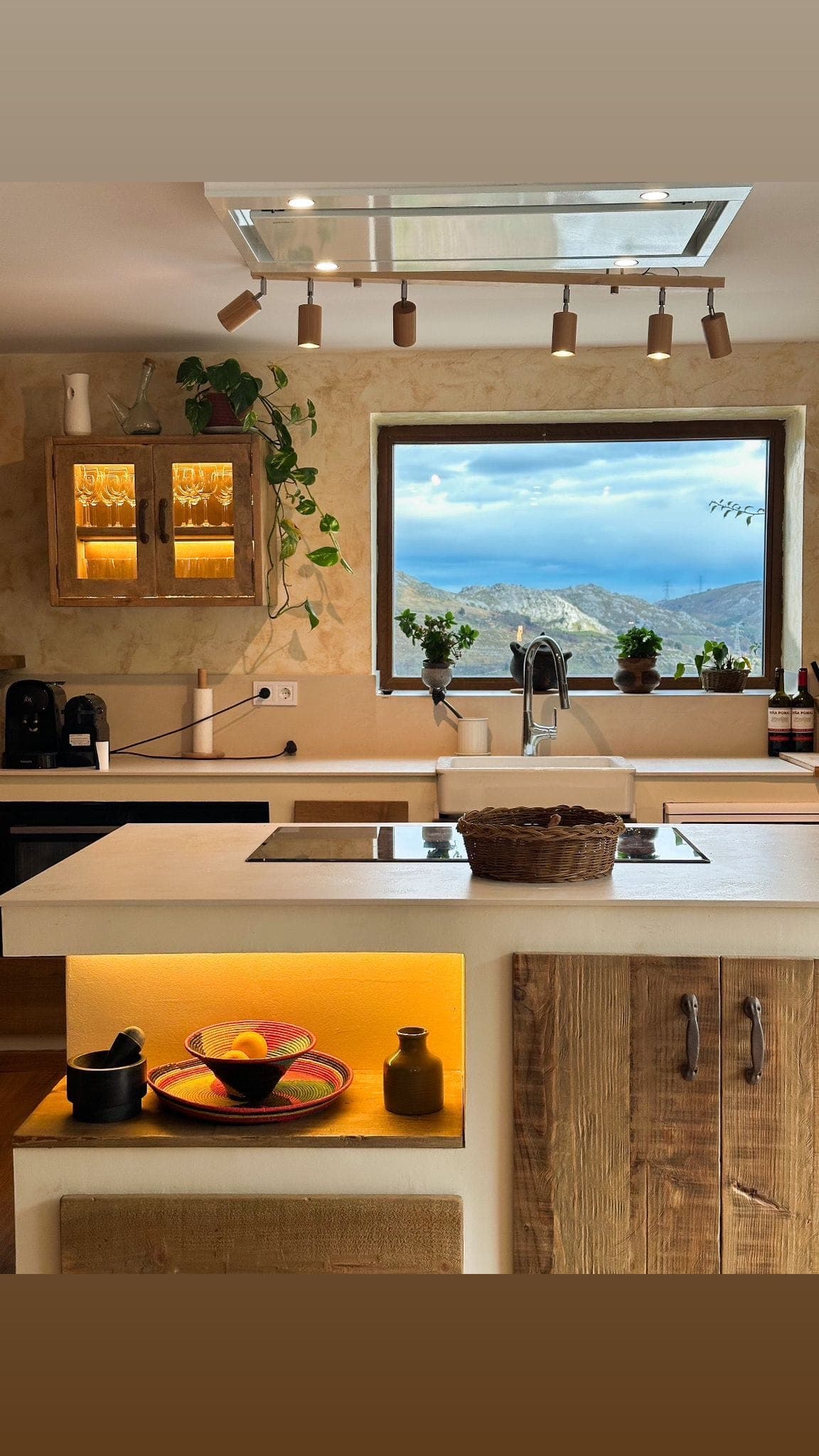A warm kitchen with a large window overlooking the mountains. A basket sits on the central island, with plants, kitchen utensils and a fruit bowl on display. Warm lighting highlights the rustic wood cabinets and worktops, creating a cosy atmosphere.