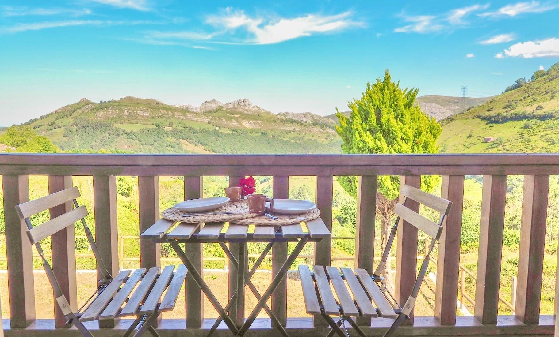 Two wooden chairs and a small table with a woven placemat, plates and cups are set on a balcony. The view overlooks a green valley, rolling hills and a clear blue sky in the background.
