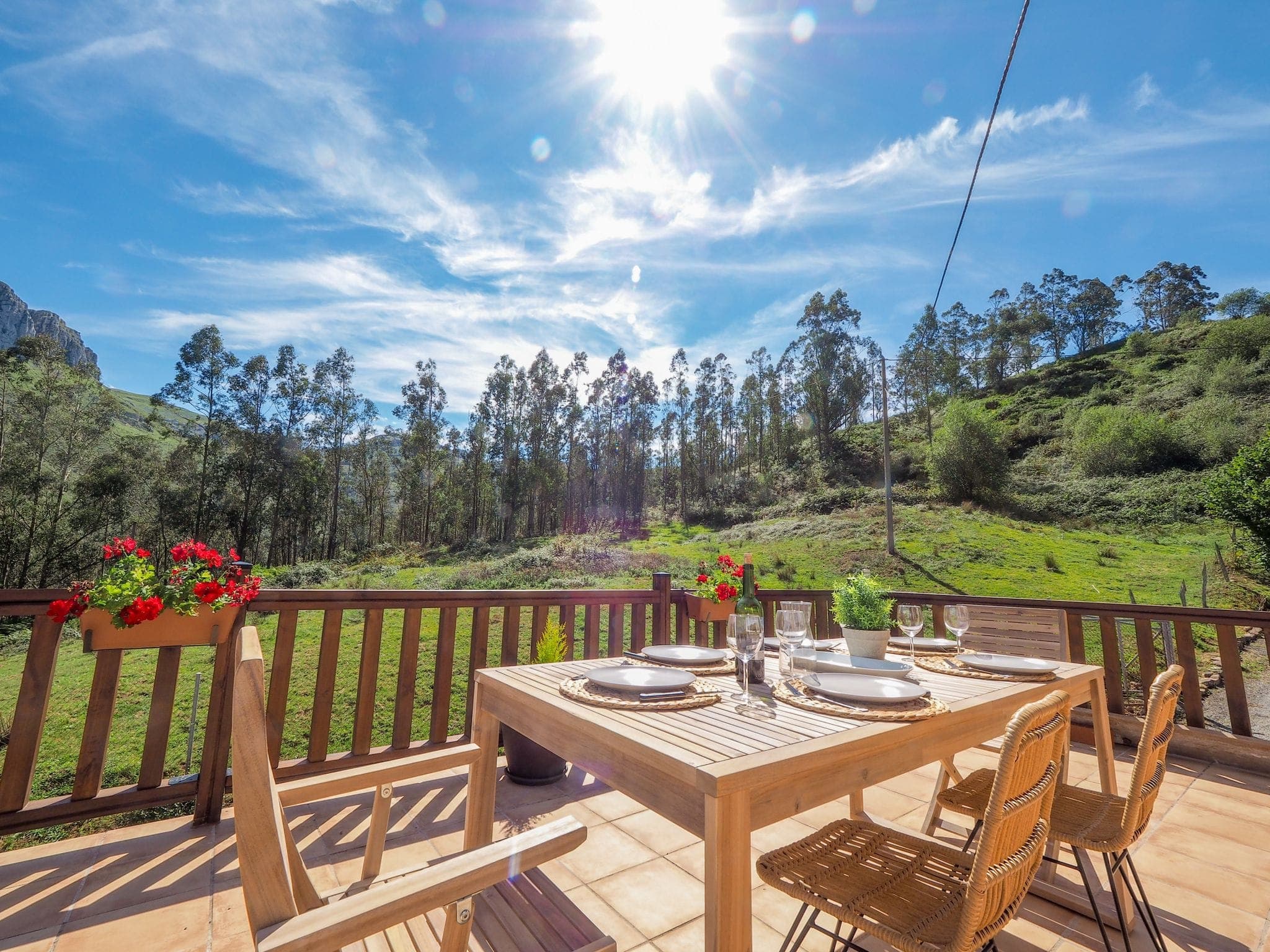 A wooden dining table, complete with plates, glasses and floral centerpiece, sits on a sunny terrace. Surrounding it, a lush green landscape of trees and hills, under a bright blue sky and radiant sunshine.
