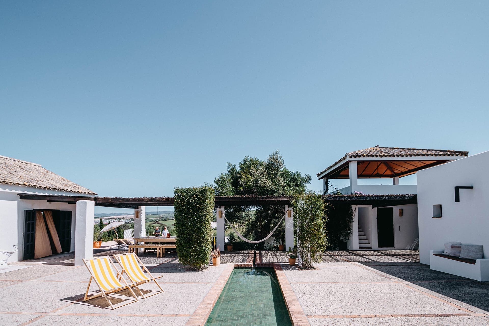 A modern outdoor patio with yellow-striped lounge chairs, a hammock between two green hedges, a narrow pool, and white buildings with tiled roofs under a clear blue sky.