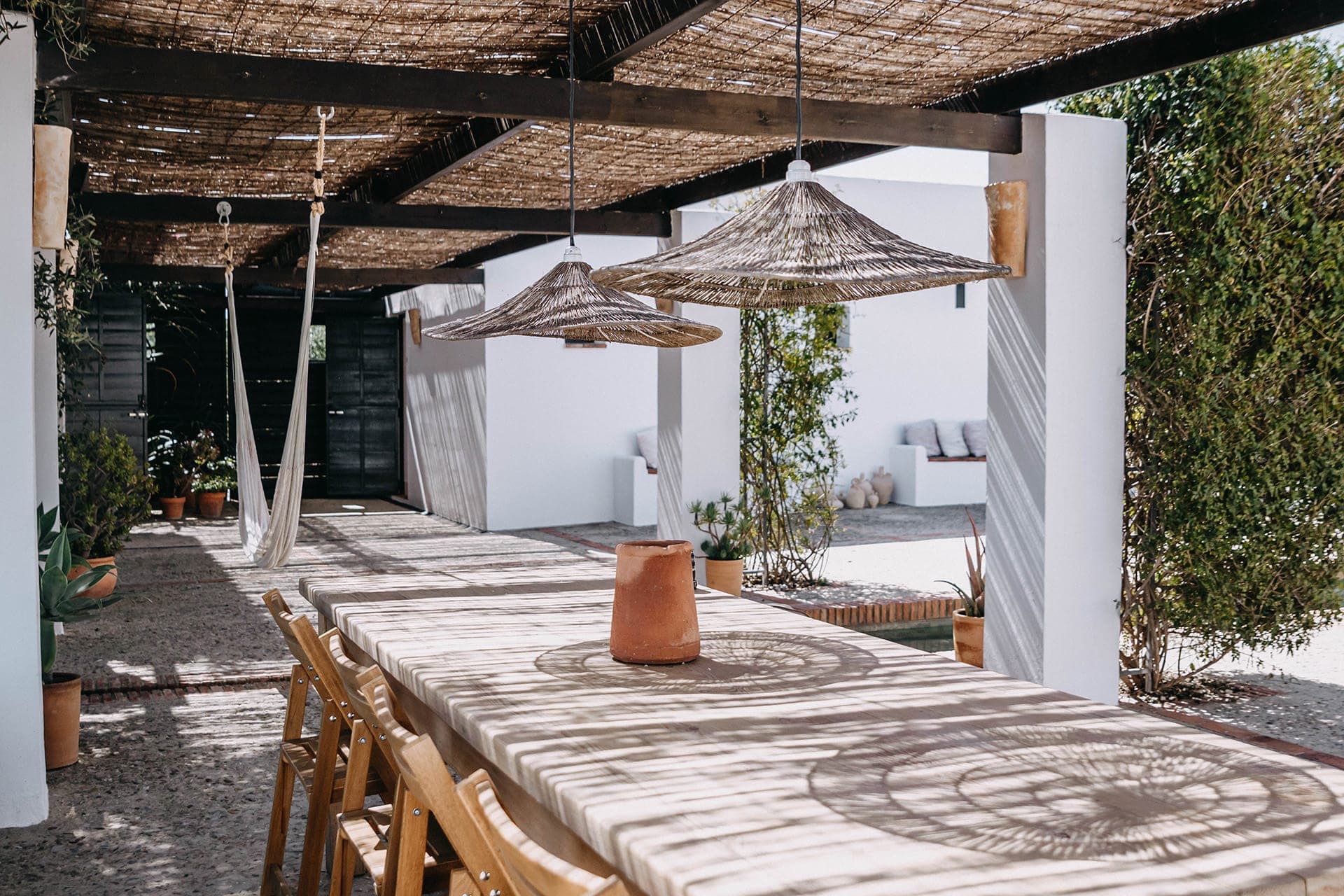 Outdoor dining area with a long wooden table and chairs under a pergola. Rattan lights hang overhead, creating soft shadows. The walls are white, surrounded by greenery, and a hammock is visible in the background. Sunlight filters through the woven roof.