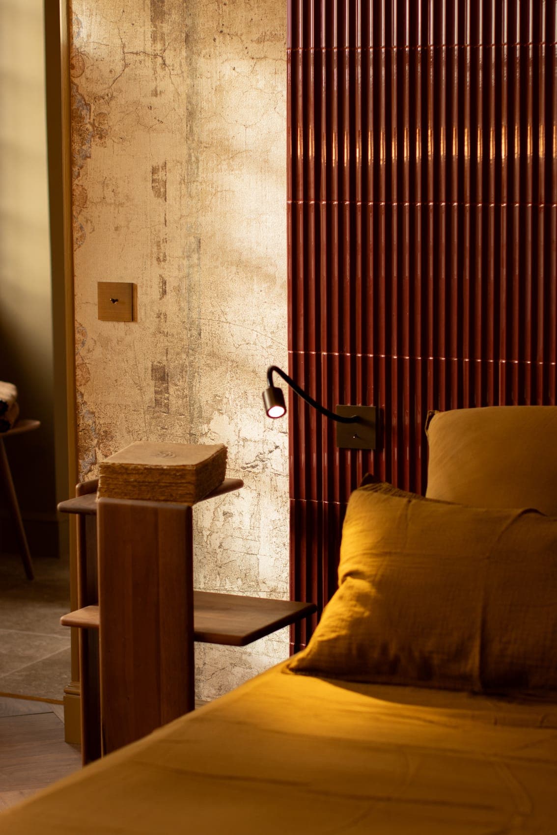 Coin chambre cosy avec un lit jaune, un mur beige texturé, un panneau à lattes verticales rouges et une petite étagère en bois contenant des paniers tressés, chaleureusement éclairé par la lumière naturelle et une lampe de lecture murale.