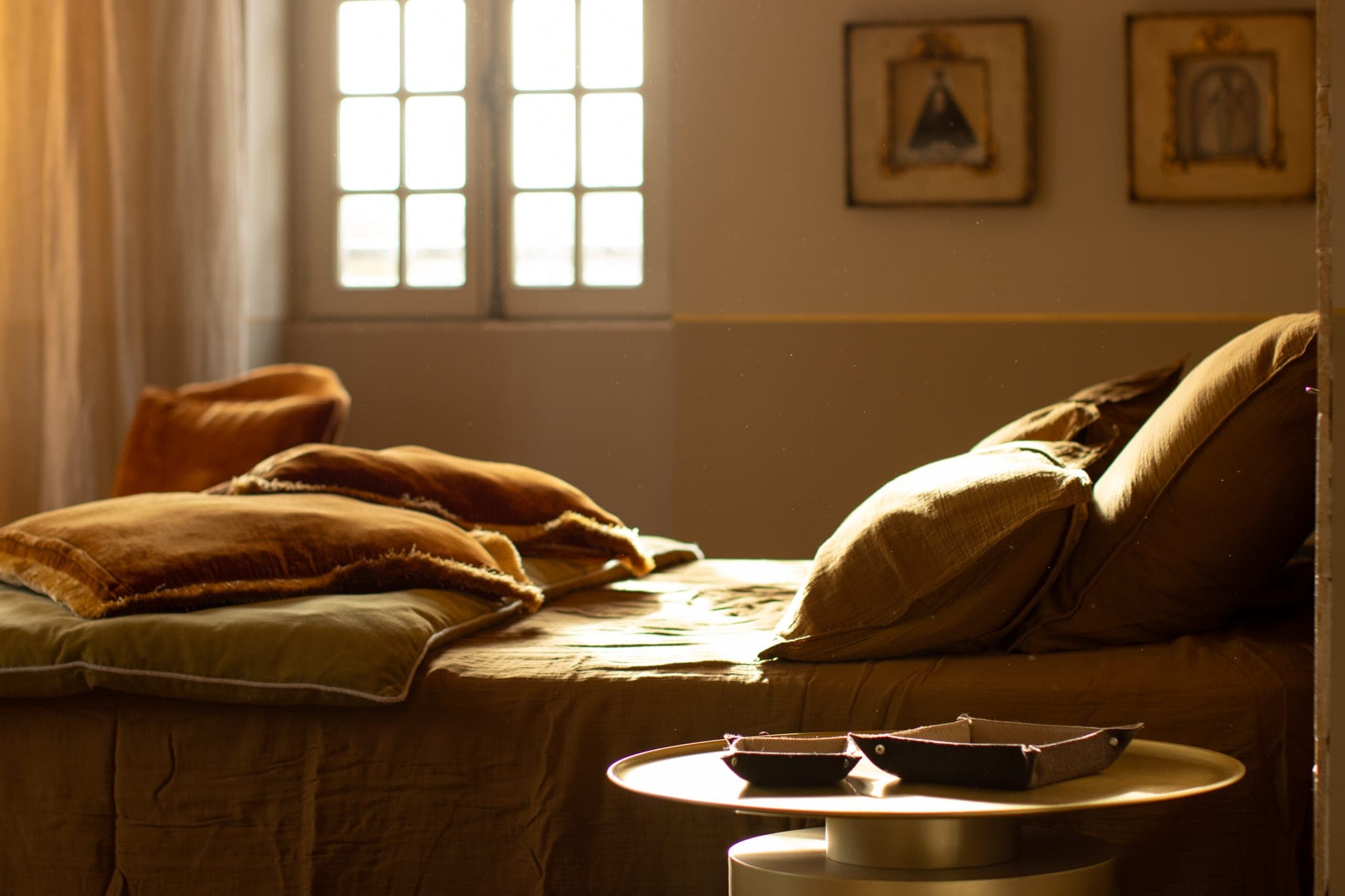 Une chambre douillette baignée d'une douce lumière dorée par la fenêtre, illuminant un lit soigneusement fait, agrémenté d'oreillers et de couvertures marron et beige. Une petite table ronde avec deux plats foncés trône au premier plan.