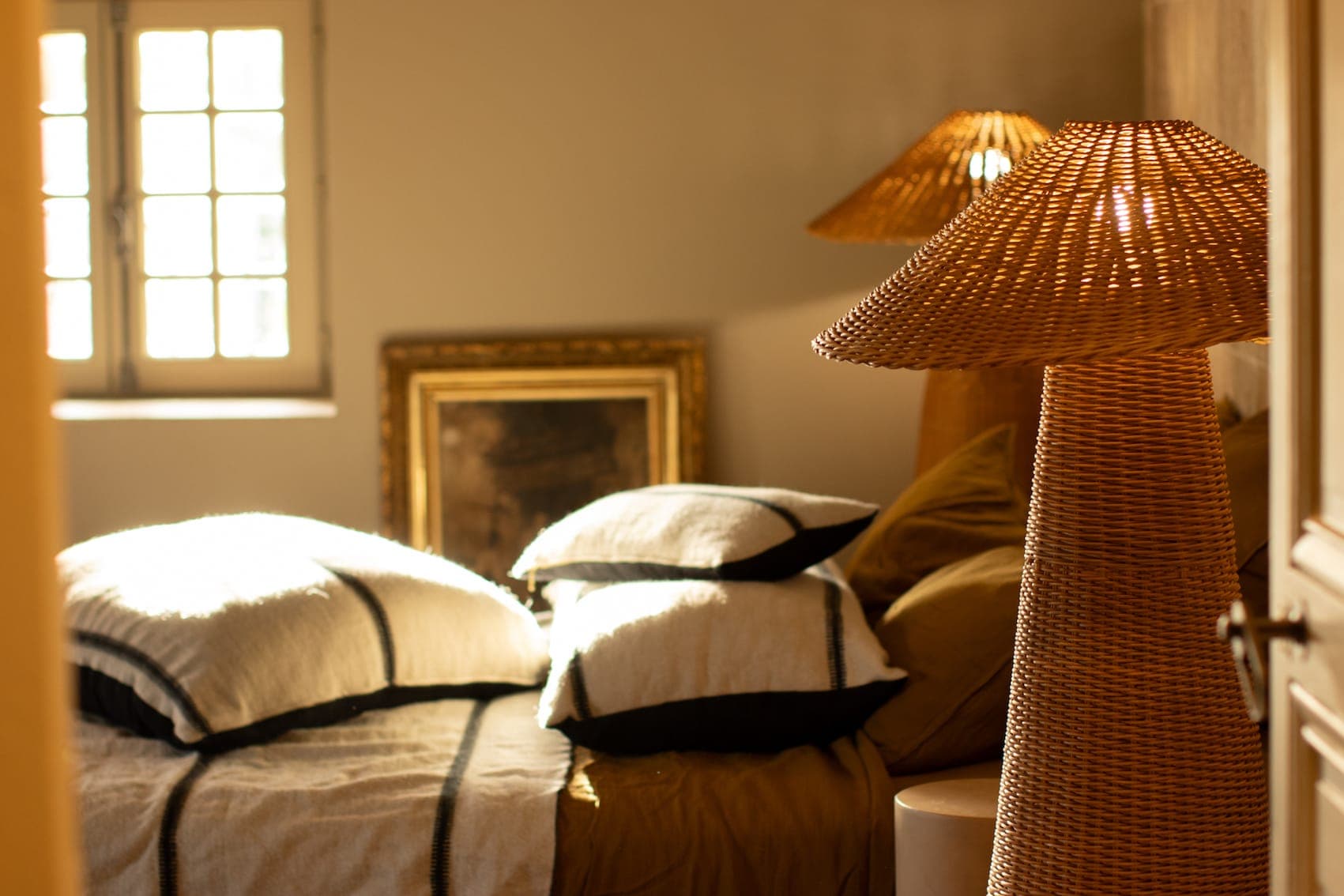 Une chambre douillette, baignée de soleil par une fenêtre, illumine un lit soigneusement fait, agrémenté d'oreillers blancs et noirs et d'une couverture marron. On y voit également deux lampes en rotin tressé et un tableau encadré.