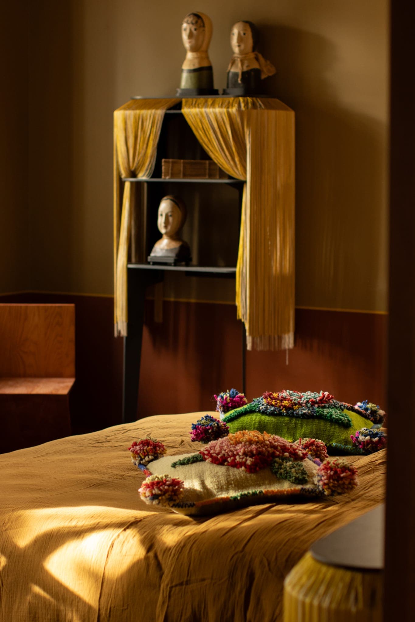 Une chambre chaleureusement éclairée avec un couvre-lit jaune moutarde, une couverture texturée colorée sur le lit et une étagère noire contenant des bustes décoratifs surmontés de rideaux à franges jaunes contre un mur beige.