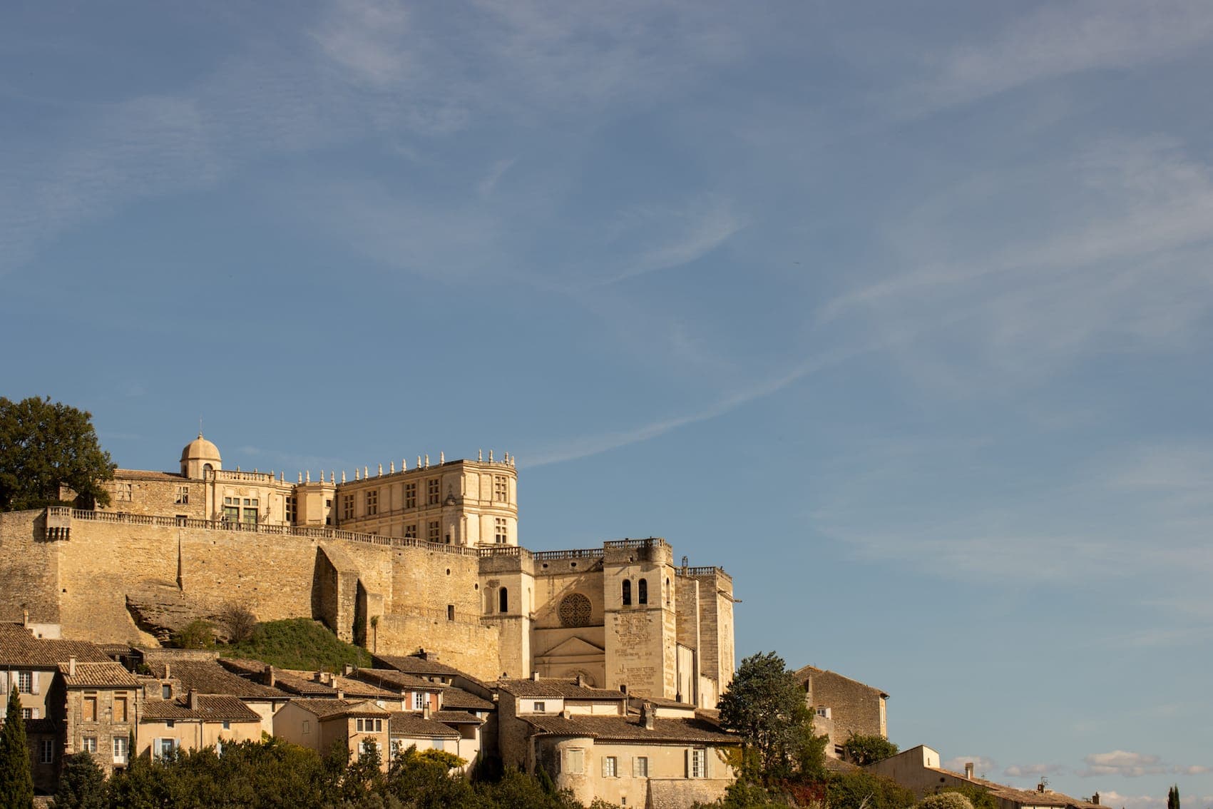 Un château historique en pierre se dresse au sommet d'une colline, surplombant un ensemble de maisons anciennes, sous un ciel bleu parsemé de nuages fins. La lumière du soleil projette des tons chauds sur les bâtiments et la verdure entoure le quartier.