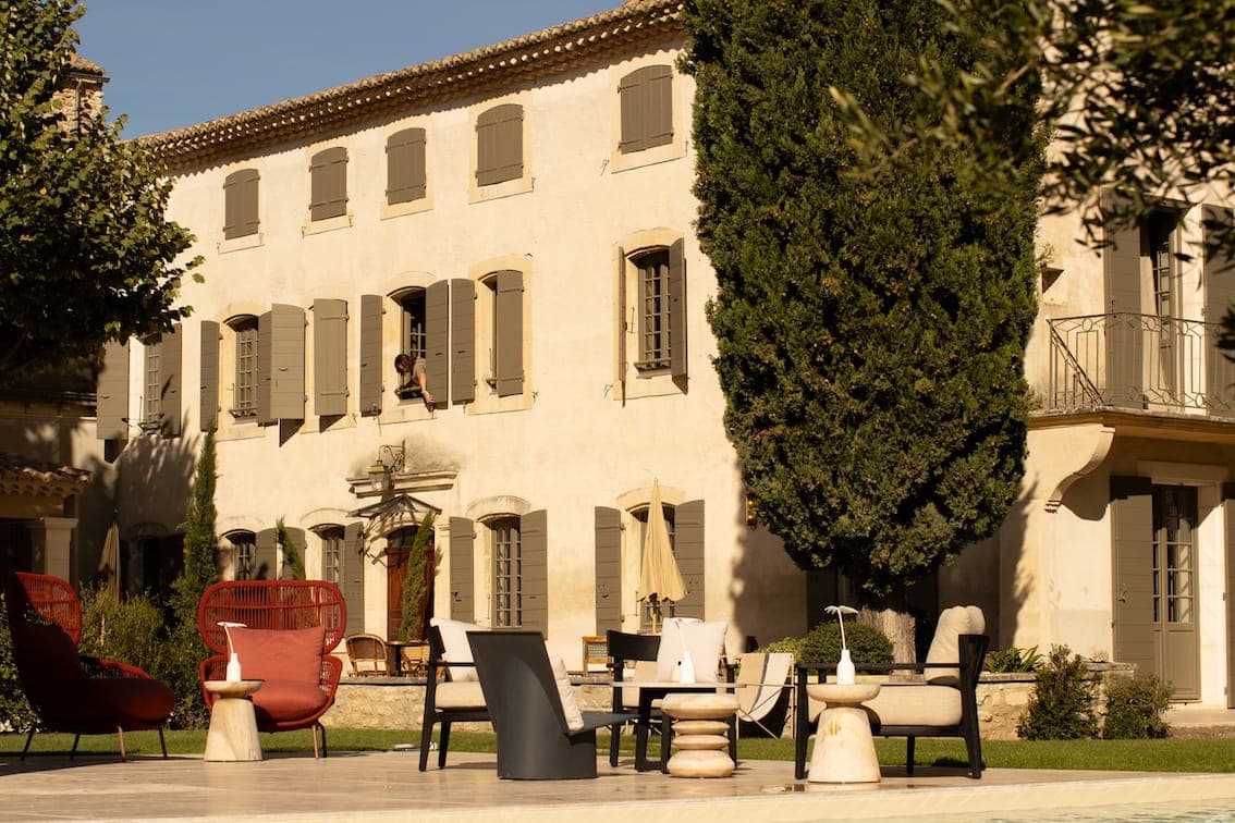 Une villa ensoleillée aux murs beiges et aux volets gris, entourée d'arbres. Un mobilier d'extérieur moderne aux tons rouge, noir et blanc est disposé près d'un patio en pierre. Une personne regarde par une fenêtre à l'étage.