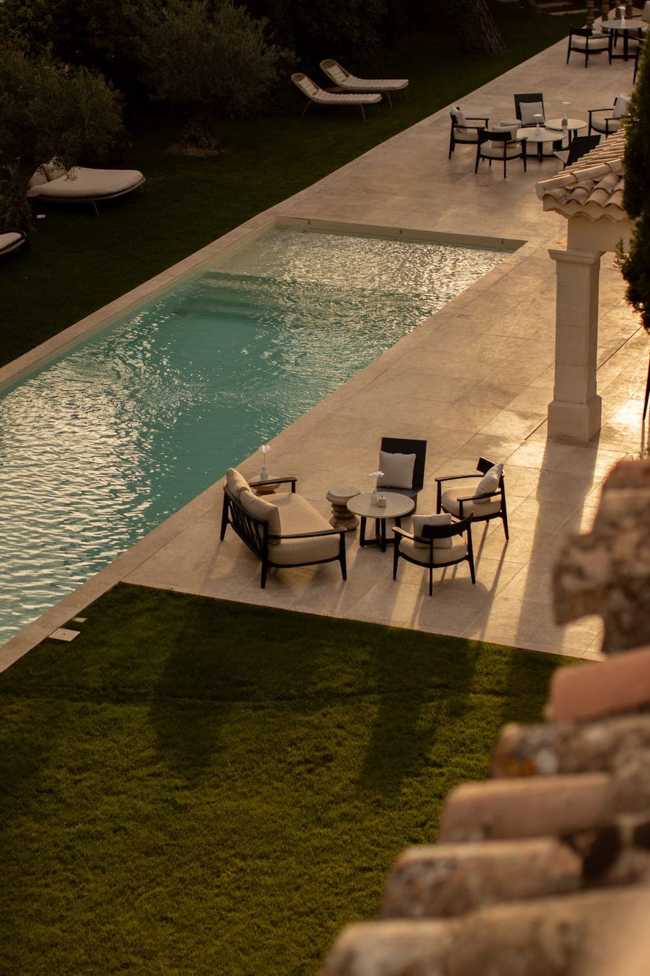 Un patio ensoleillé à côté d'une longue piscine comprend des chaises longues et des tables rembourrées, avec d'autres sièges et chaises longues dispersés le long de la terrasse de la piscine au milieu de l'herbe verte et des arbres.
