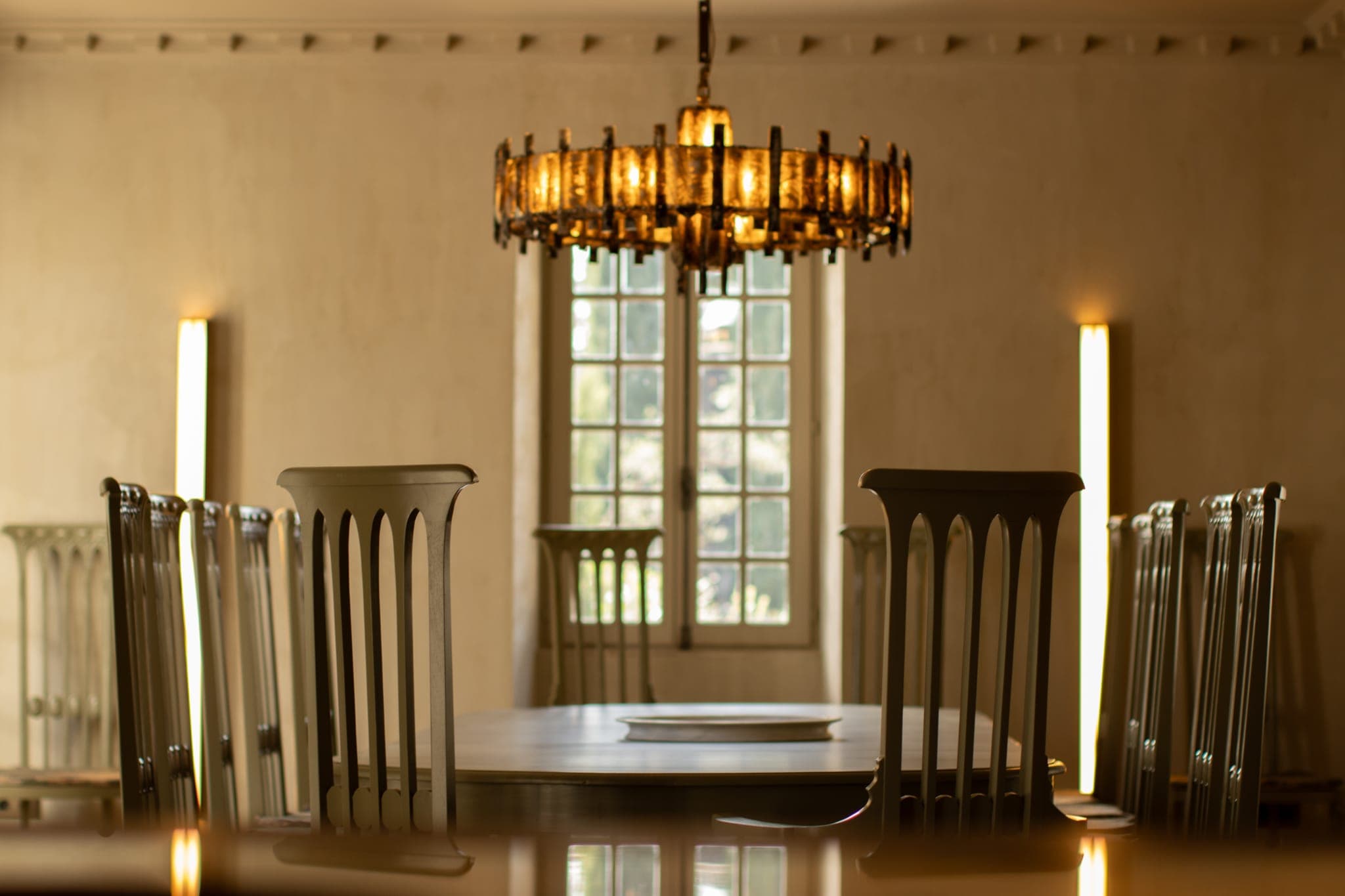 Une salle à manger avec des chaises en bois à haut dossier autour d'une table, sous un grand lustre doré orné. La lumière du soleil filtre à travers une fenêtre en arrière-plan, projetant une douce lueur dans la pièce.