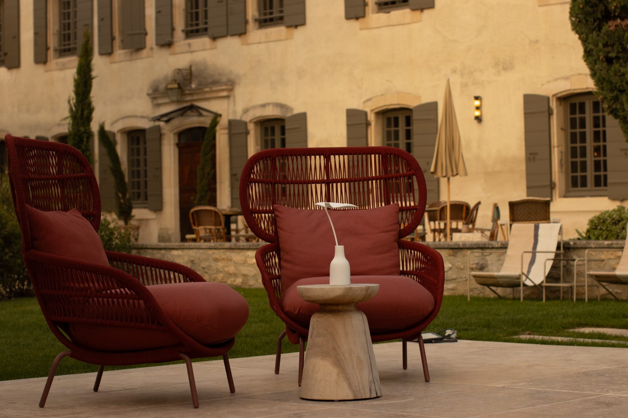 Deux chaises d'extérieur rouges modernes avec coussins sont assises près d'une petite table ronde contenant un vase blanc, installée sur une terrasse devant un élégant bâtiment beige avec des fenêtres à volets et un jardin bien entretenu.