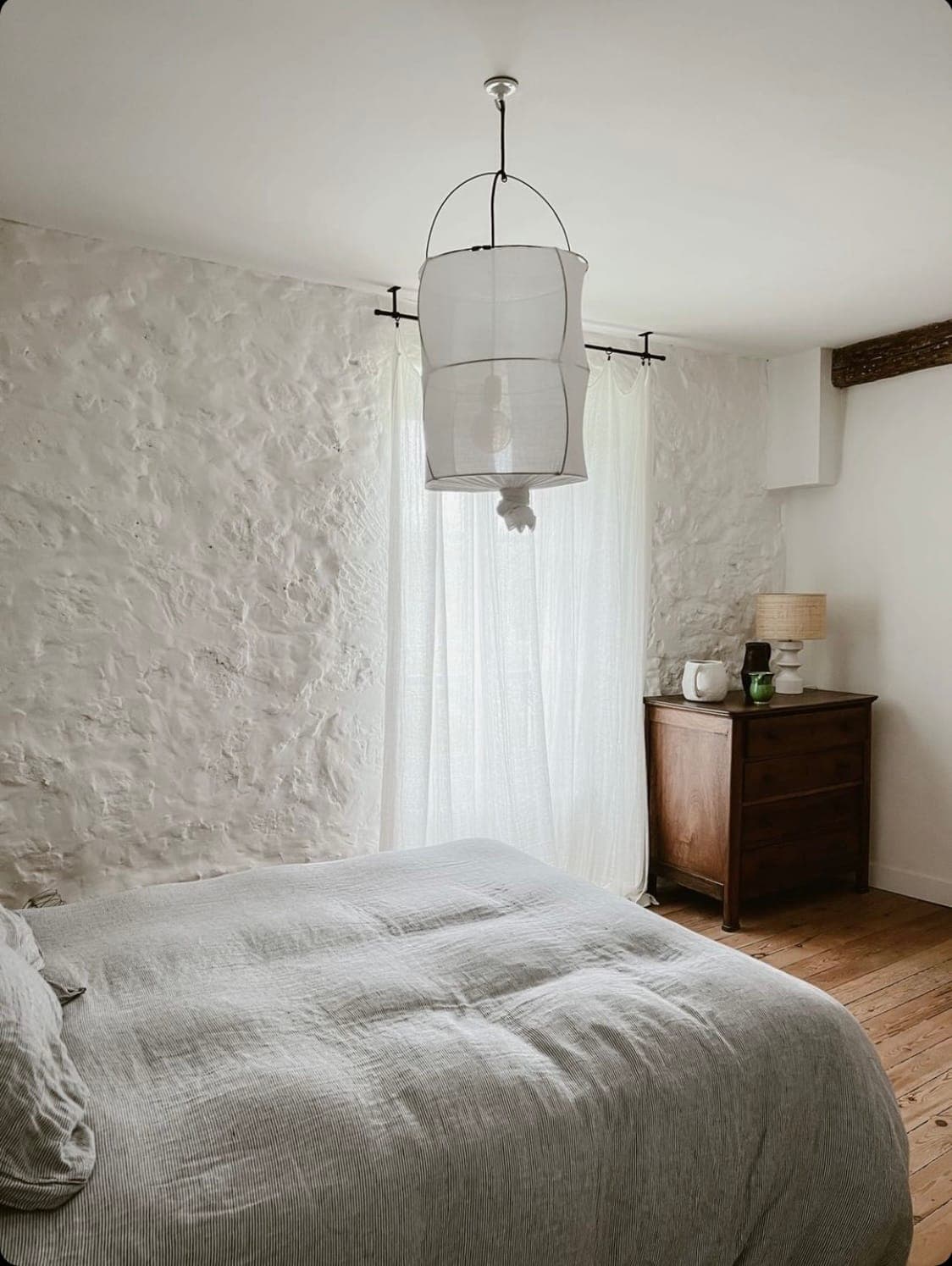 A cosy bedroom with a textured white wall, a bed covered with a gray blanket, a wooden chest of drawers and a hanging lamp. Transparent curtains gently diffuse natural light from the window, creating a serene atmosphere.