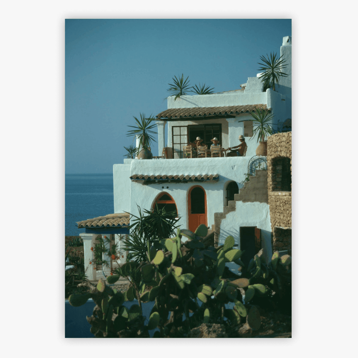 Villa blanche de style méditerranéen surplombant la mer, avec toit en tuiles de terre cuite et fenêtres en arc arrondi. Des palmiers ornent les terrasses. Le ciel bleu éclatant et la mer calme créent une atmosphère sereine.